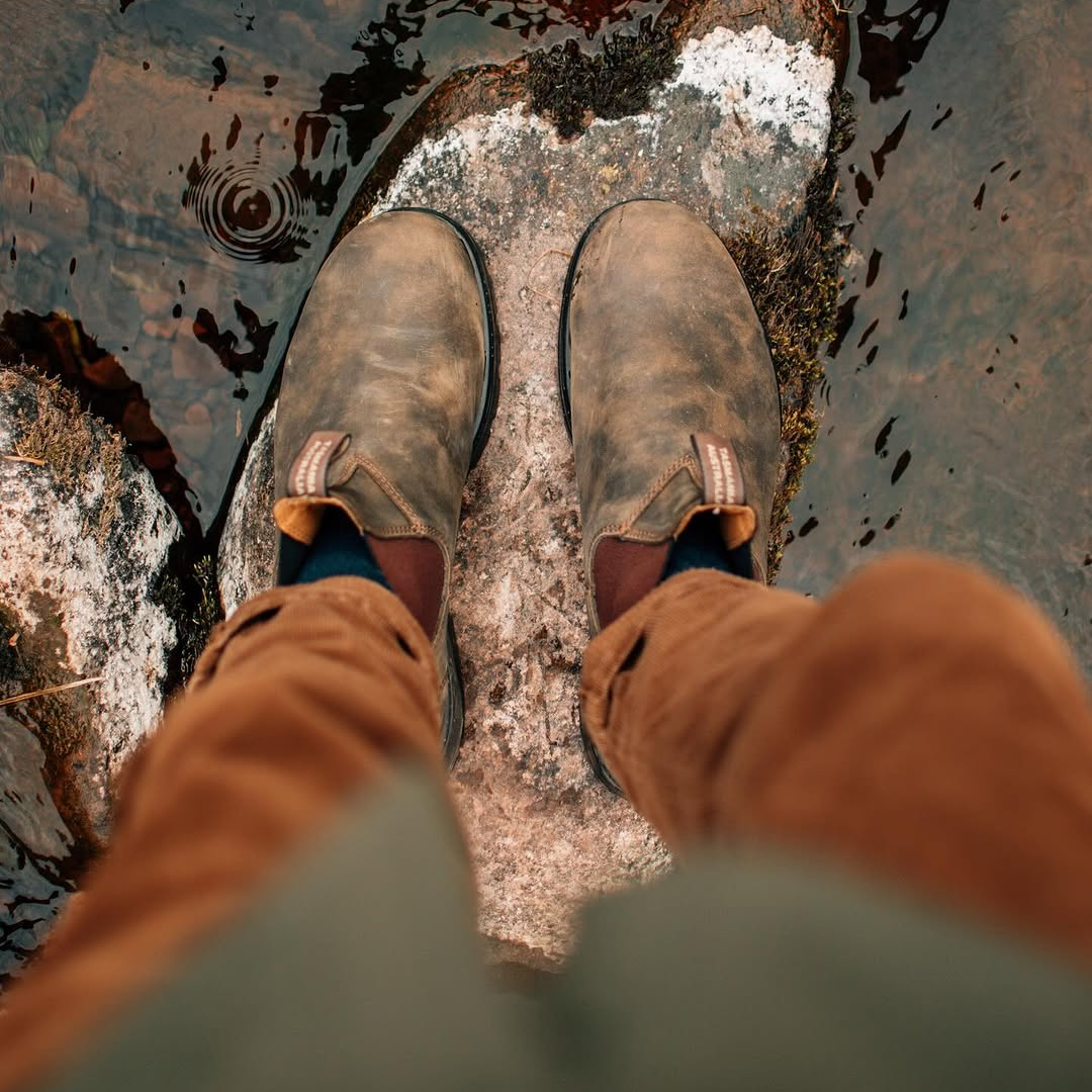 Photo from above showing a person standing on a large rock at the edge of a body of water, wearing brown boots and brown pants.