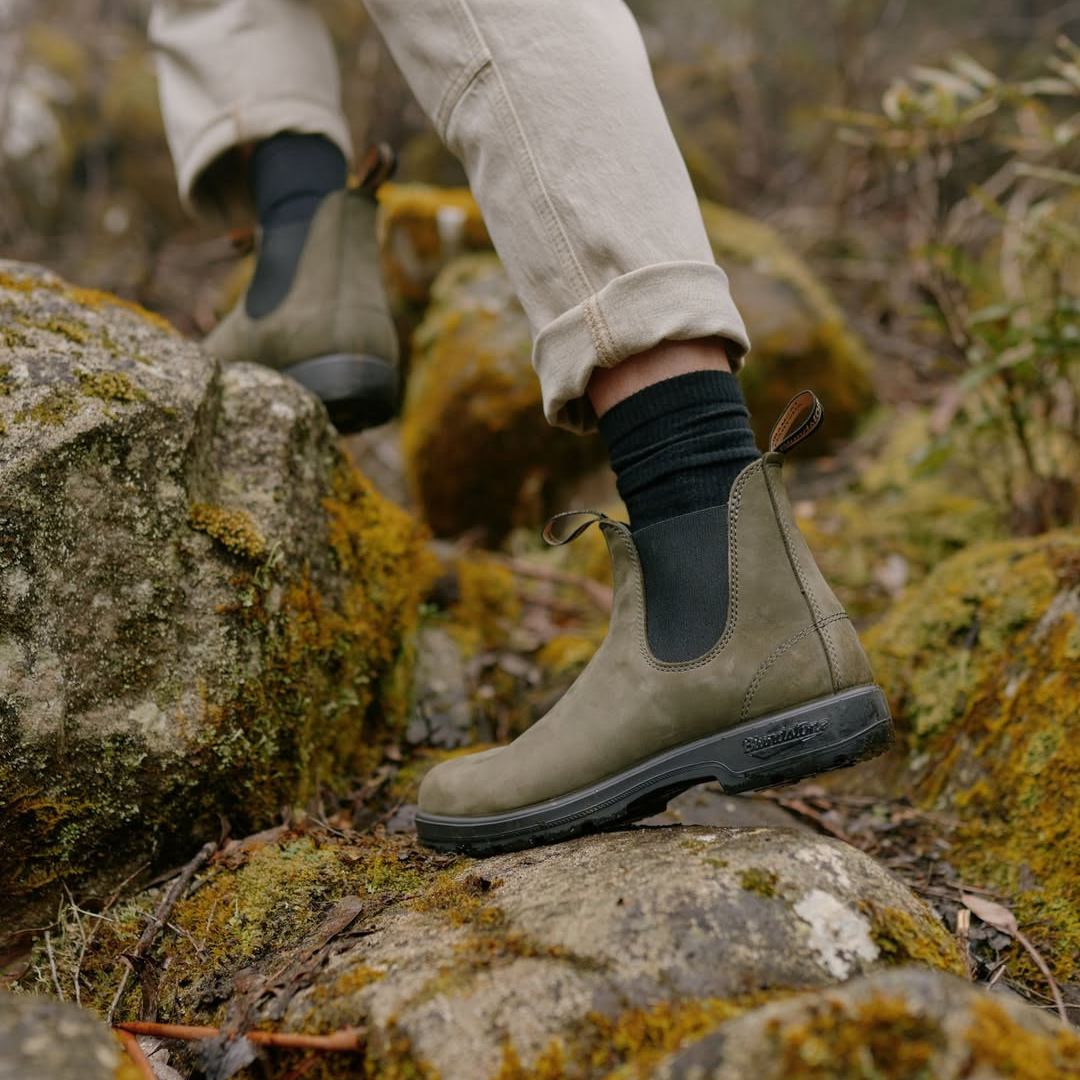 Person wearing beige pants, black socks, and waterproof hiking boots stepping on moss-covered rocks outdoors.