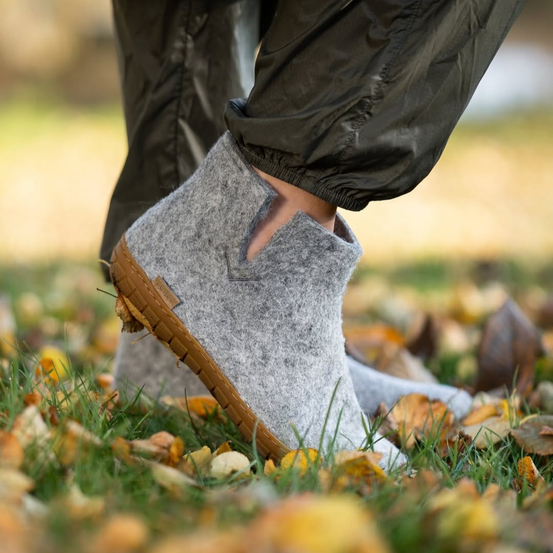 Close-up of a person's ankle wearing a grey felt ankle boot with a brown sole, walking on fallen autumn leaves in a park.