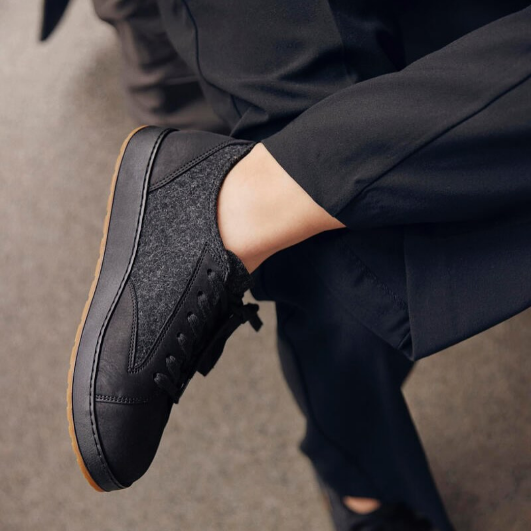Person wearing black shoes with gray felt panels, black pants, and black shirt, sitting with their legs crossed.