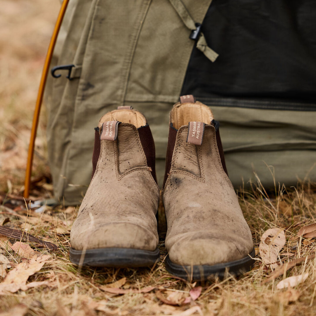 Dirty brown work boots placed in front of a green tent on dry grass.