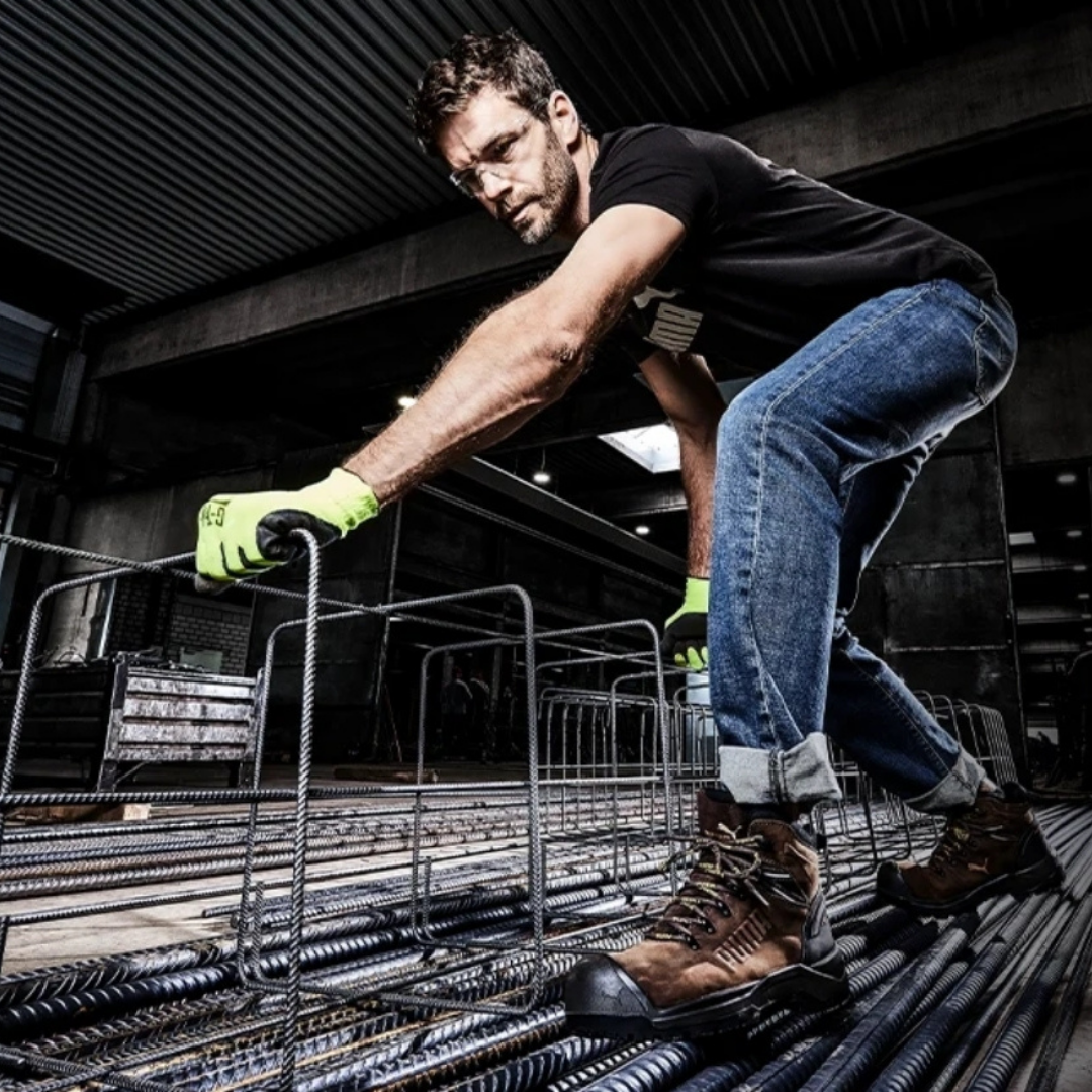 A man wearing safety glasses, gloves, jeans, and work boots is working on a construction site, handling steel rebar.