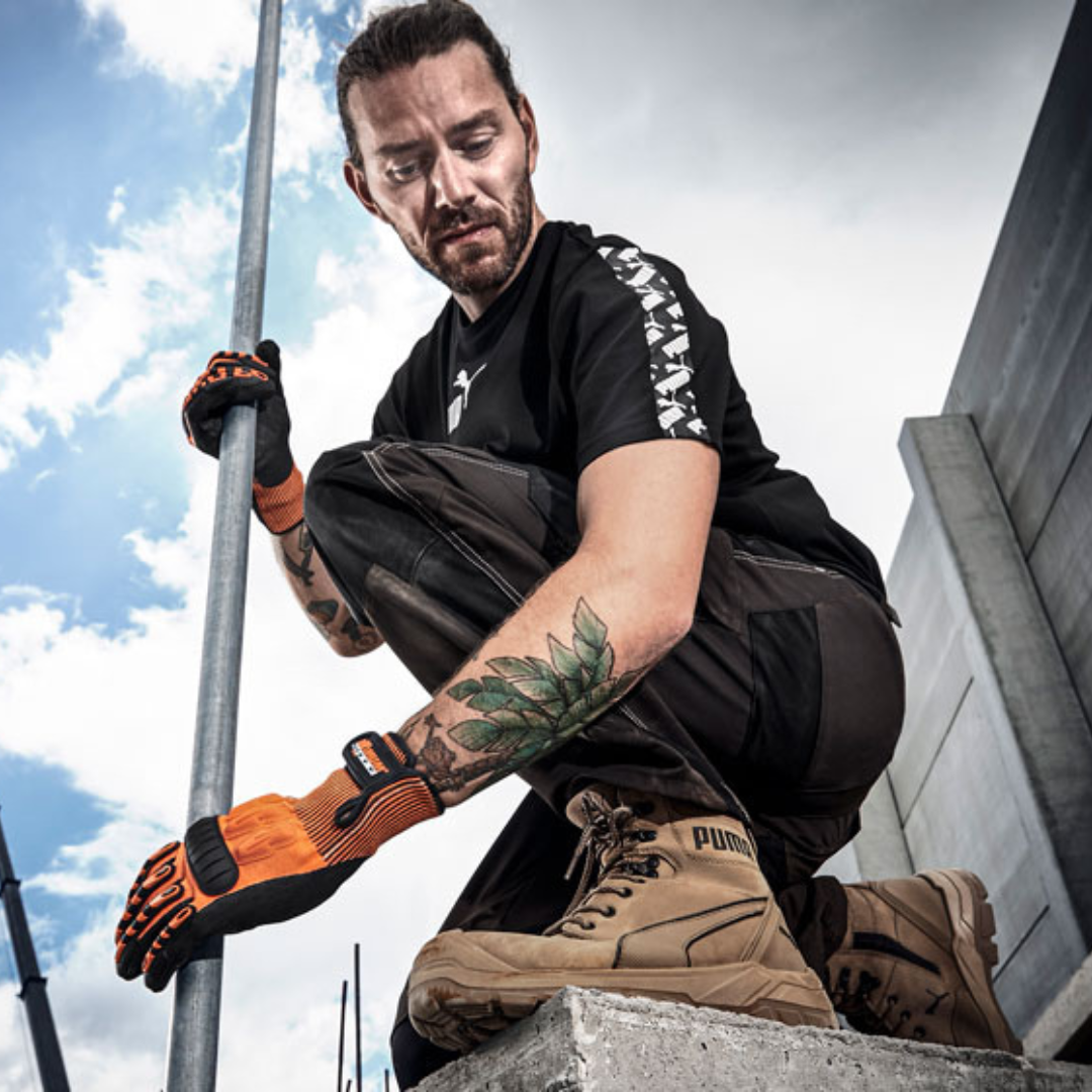 A man wearing beige work boots, black work pants, and a black T-shirt with a pattern on the sleeves, is crouching near a concrete surface, holding a metal rod, with a cloudy sky in the background.