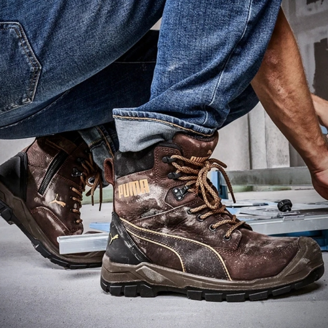 Close-up of a person wearing dirty brown Puma work boots with jeans, crouching on a workbench in a workshop.