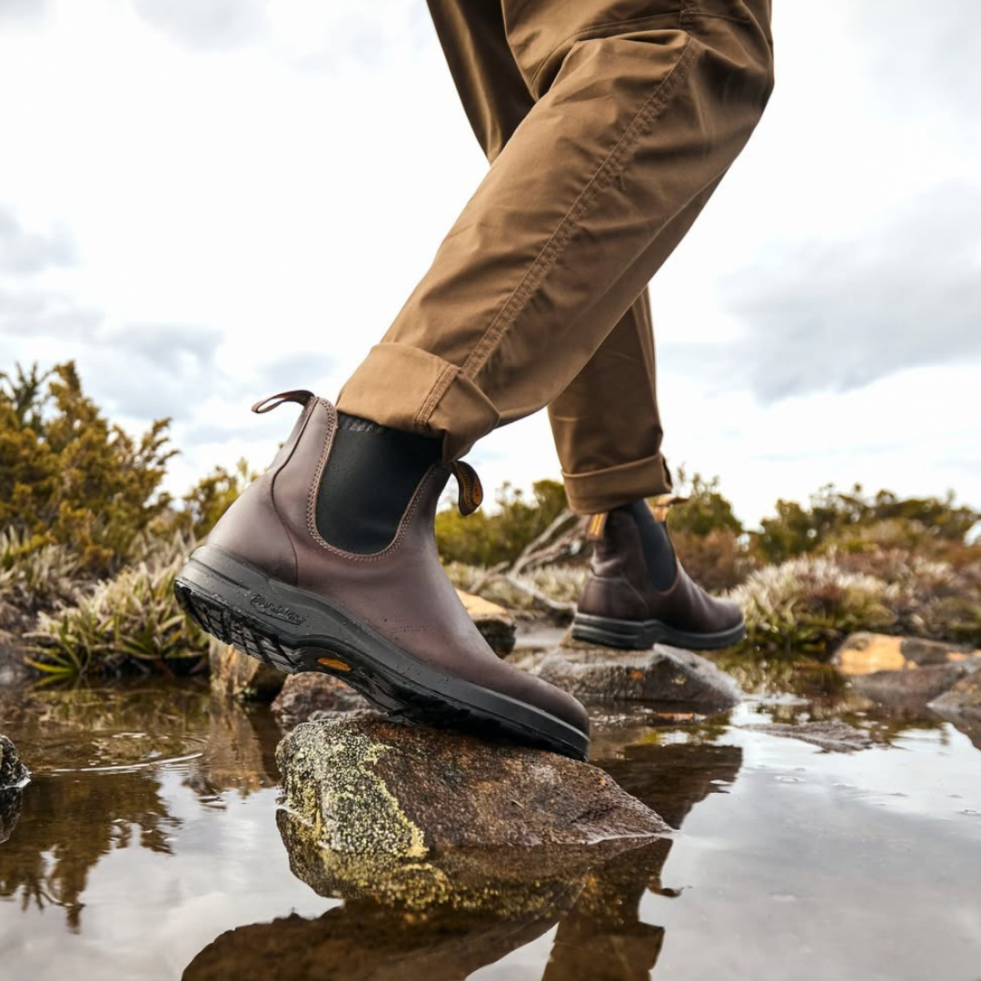 Person wearing brown waterproof boots and brown pants walking over rocks in a shallow stream, outdoor setting with cloudy sky and green shrubbery.