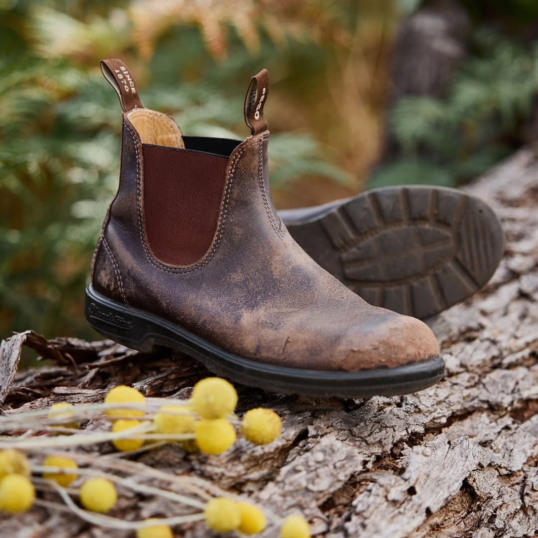 A rugged brown leather work boot placed on a fallen tree trunk with yellow flower buds nearby, outdoors with blurred green foliage in the background.