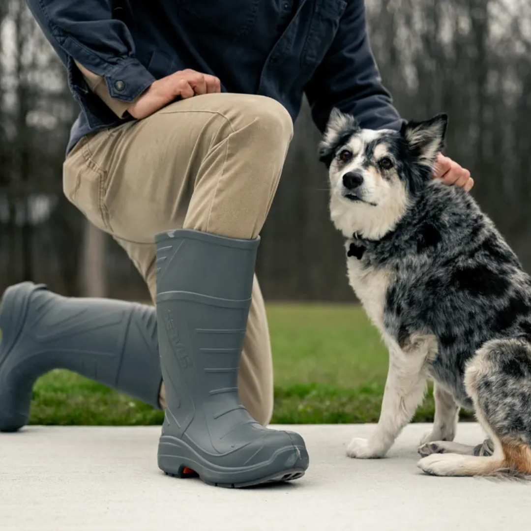 Person kneeling outdoors, wearing tan pants, a dark jacket, and gray rubber boots, with a black and white Australian Shepherd dog sitting beside them.
