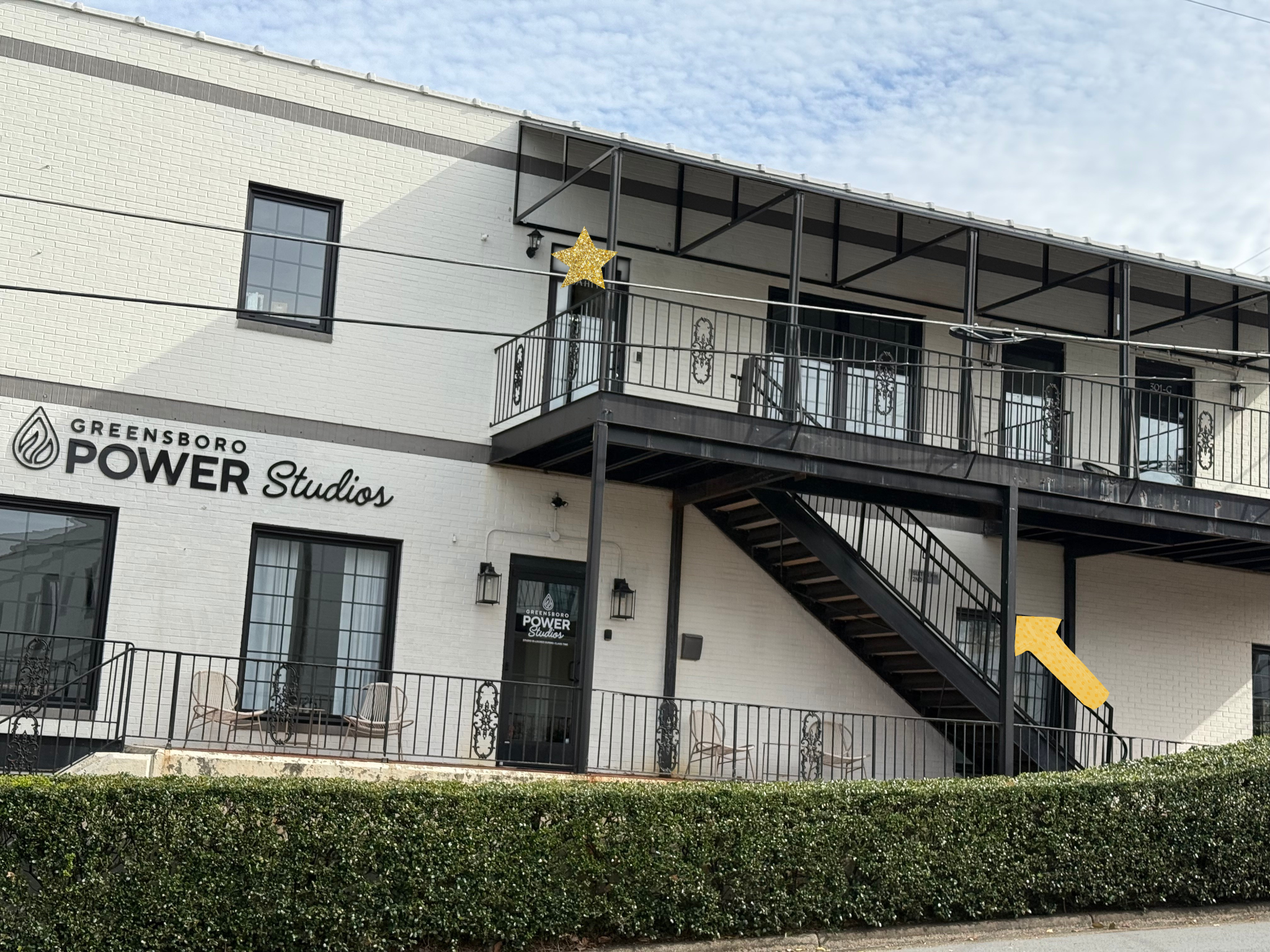 A two-story white brick building labeled 'Greensboro Power Studios' with outdoor seating, black staircase, and balcony. There is a star and arrow as directional indicators on door and staircase.