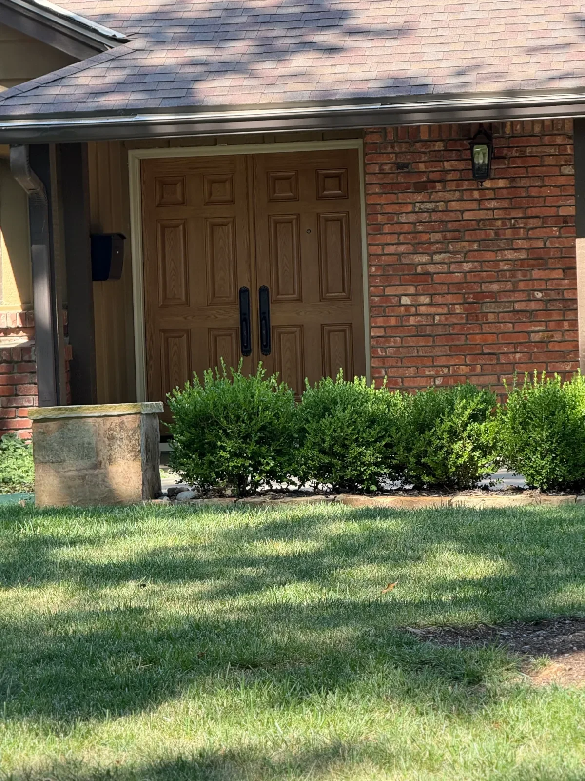 Front door of a house with a brick wall and wooden double doors, surrounded by green bushes and lawn.