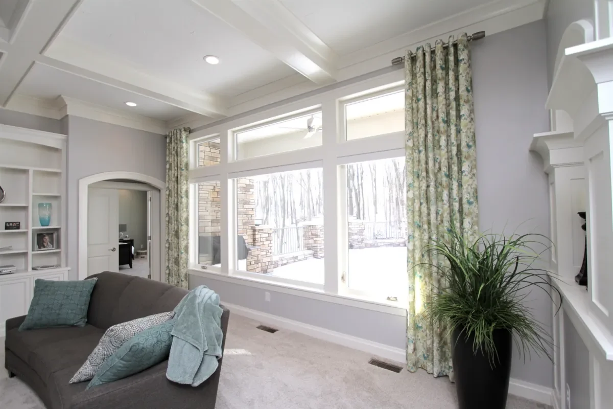 Living room with large windows, gray sofa with blue and gray pillows, patterned curtains, white built-in shelves, and a potted plant in a black vase.