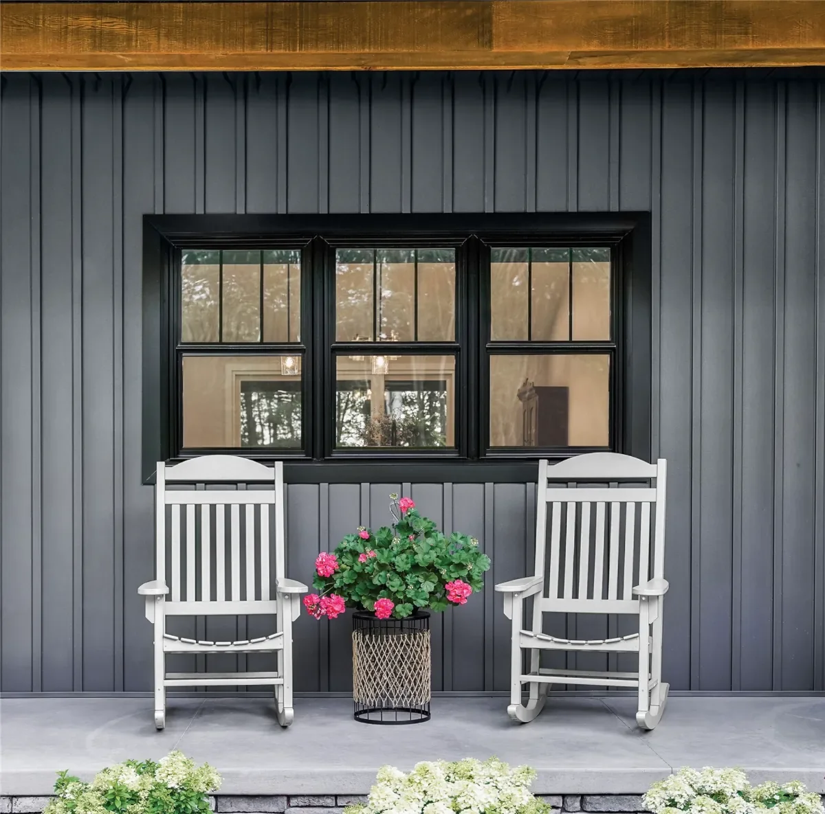 Two white wooden chairs on a porch with a black paneled wall, a large window, a pink flowering plant in a black wicker basket, and greenery at the bottom.