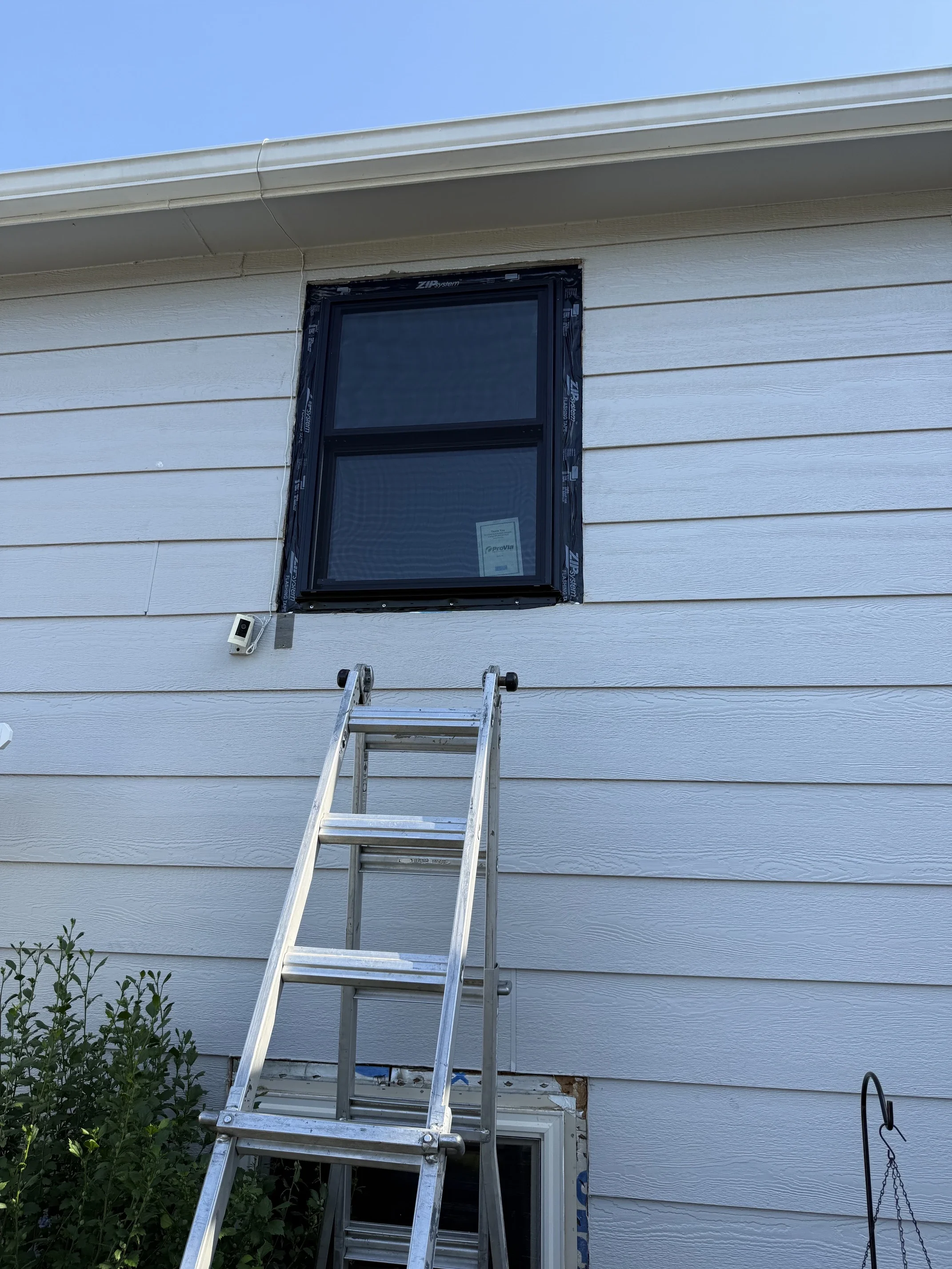 A house exterior with a window being installed or repaired, a ladder in front, bushes on the left, a small white device mounted on the wall, and a hook or tool hanging on the right side.
