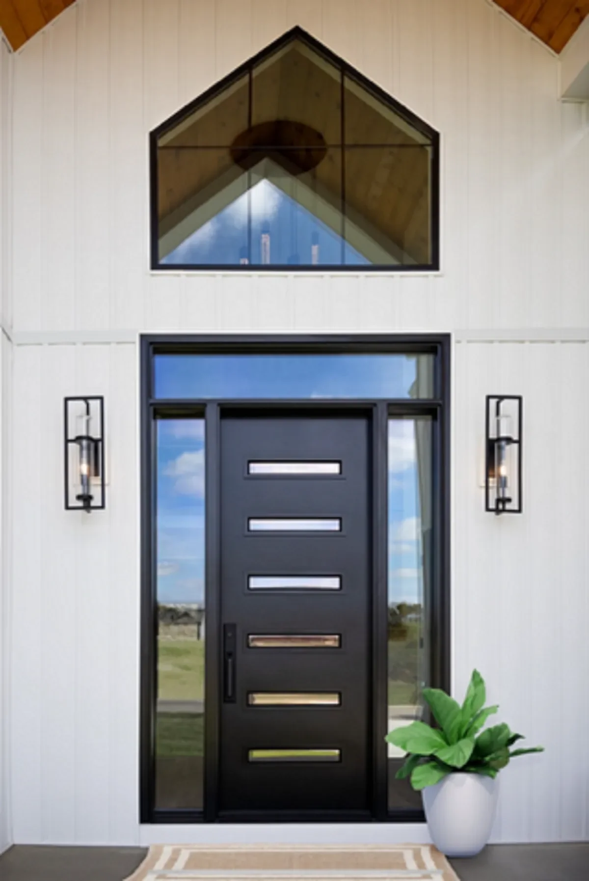 Modern front door with horizontal rectangular windows, flanked by two wall-mounted lights, and a large window above it, with a potted plant on the right and a striped rug on the porch.