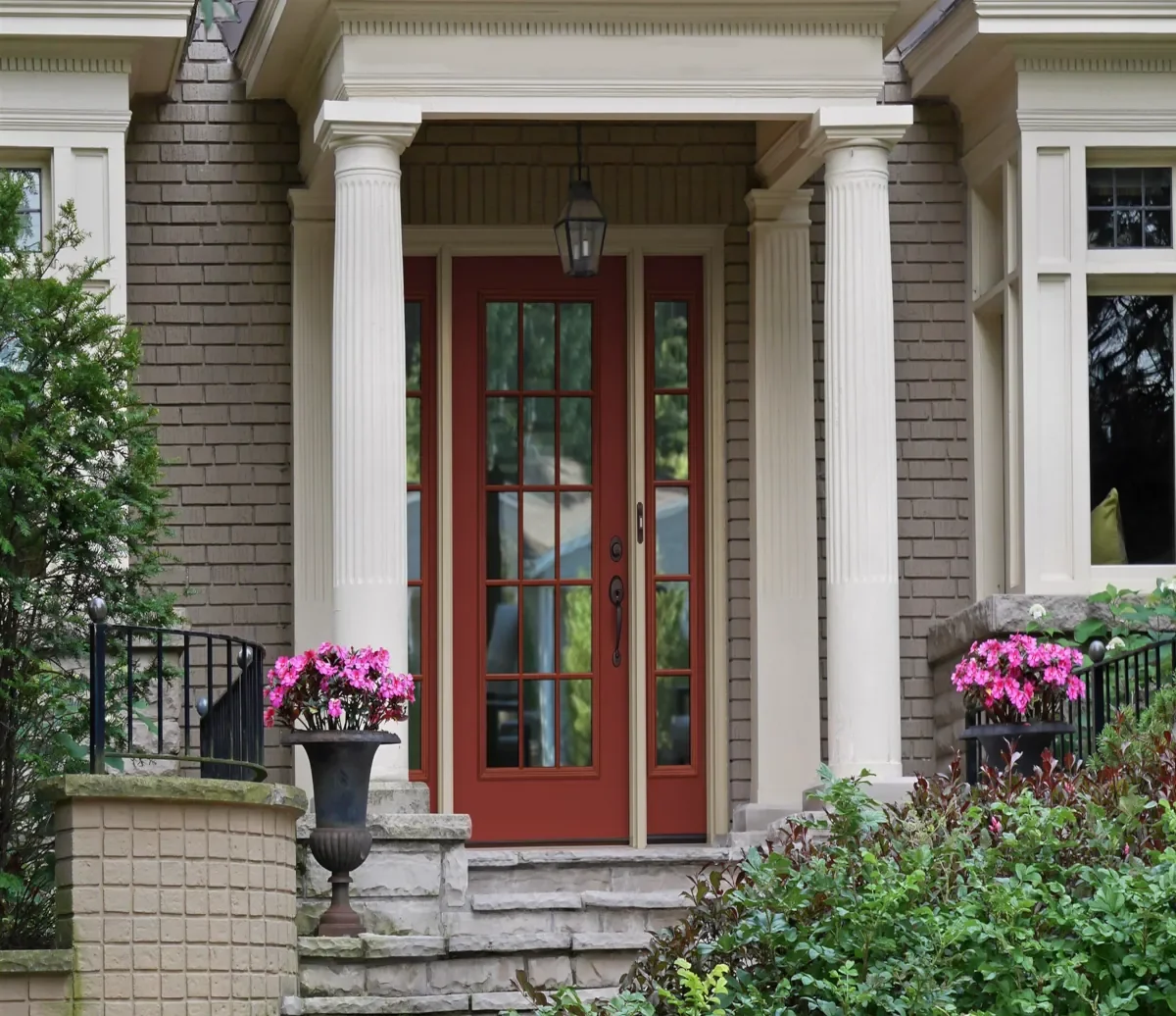 Front entrance of a house with a red door, two white columns, and potted pink flowers on either side of the steps.