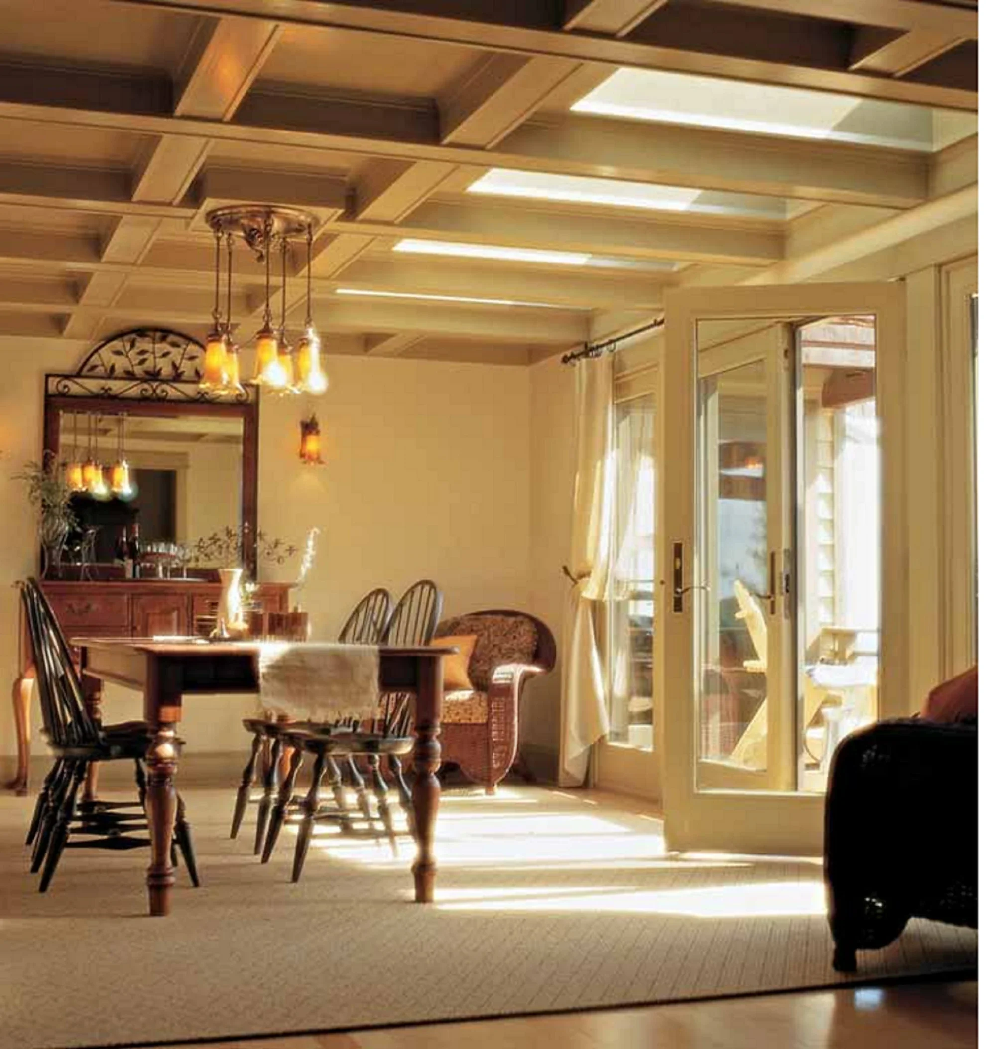 View of a dining room with a wooden table, black chairs, a wooden bench, a large mirror on the wall, a chandelier with multiple bulbs, large windows with curtains, and sunlight illuminating the room.