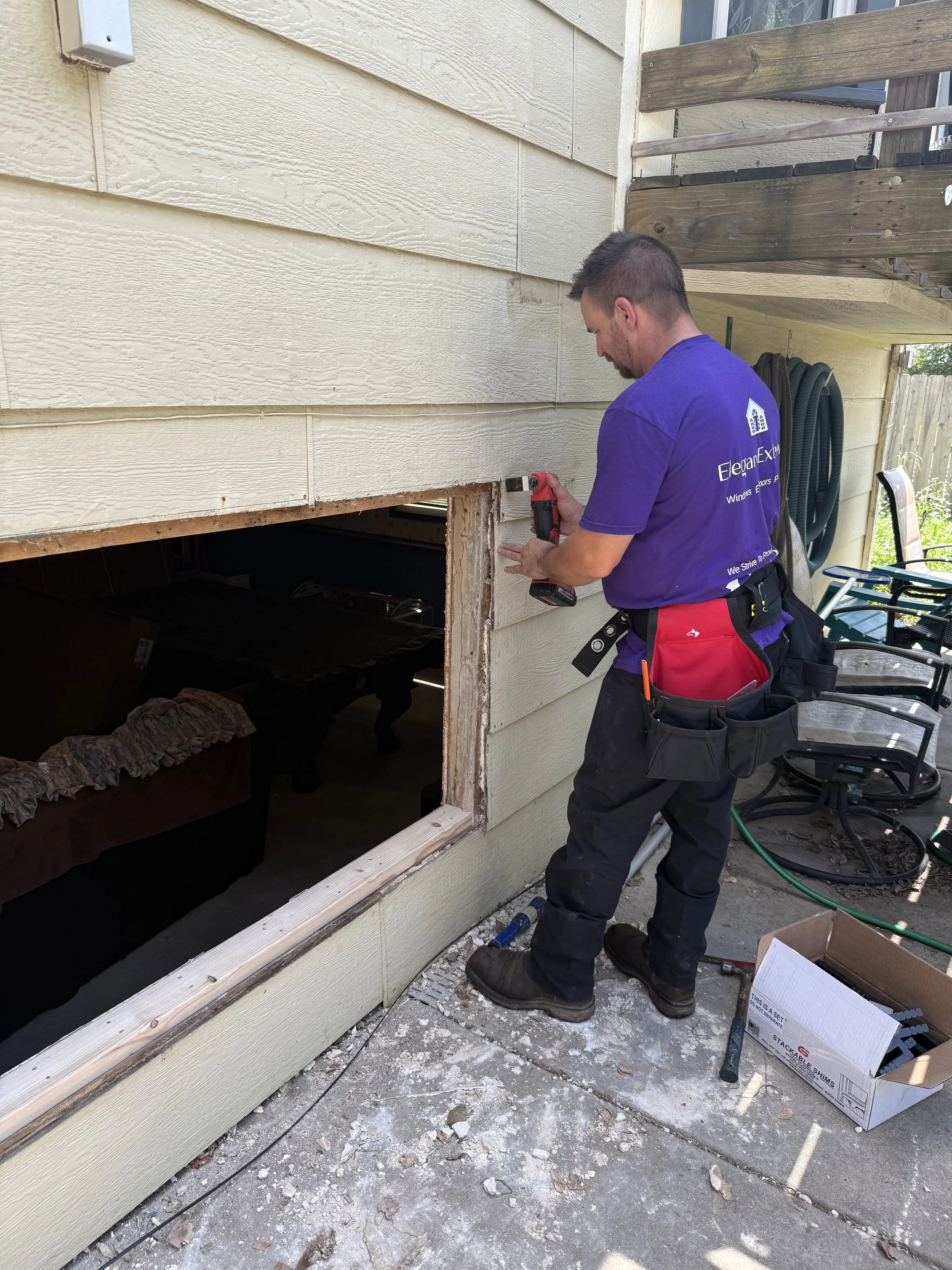 A man in a purple shirt is using a power drill on the exterior of a house, working on a window opening. The house has beige horizontal siding, and the man has a tool belt with tools around his waist. The area around him has construction debris and an open cardboard box.
