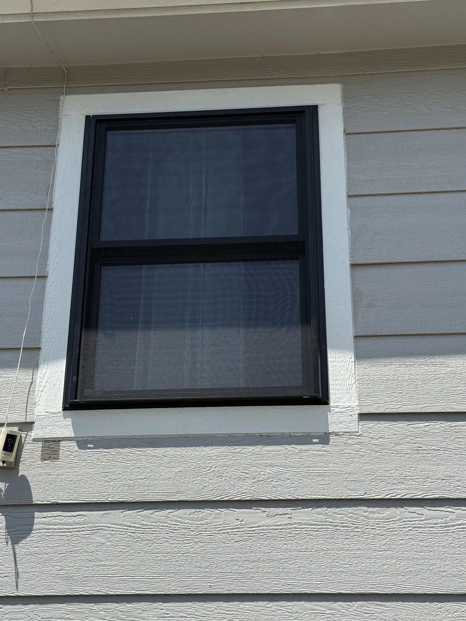 Exterior view of a house with a double-pane window, beige horizontal siding, white trim around the window, a small black security camera, and a visible power cable.