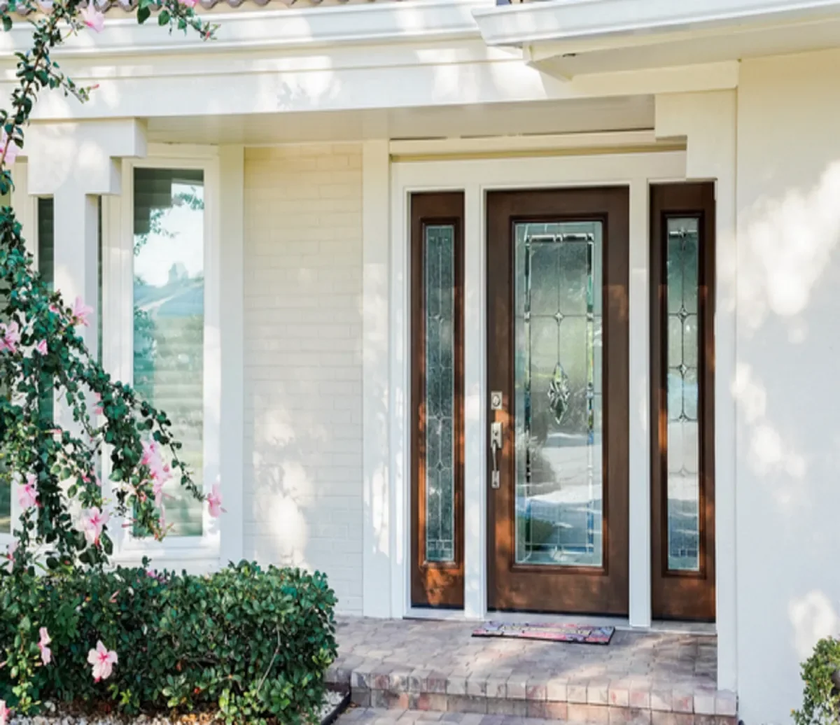 Front entrance of a house with a wooden door with glass panels, flanked by two tall narrow glass panels, and a small porch with brick pavement surrounded by green bushes and pink flowers.
