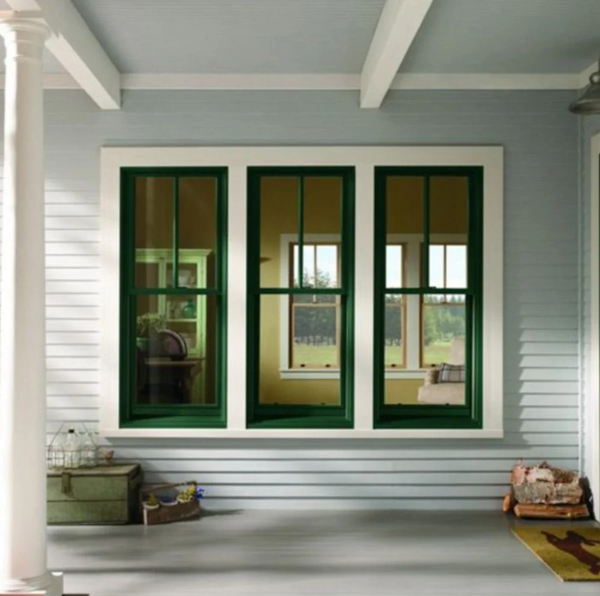 Front porch of a house with three green-framed windows, gray siding, and a white trim. Inside, a chair and bouquets of flowers are visible through the windows.