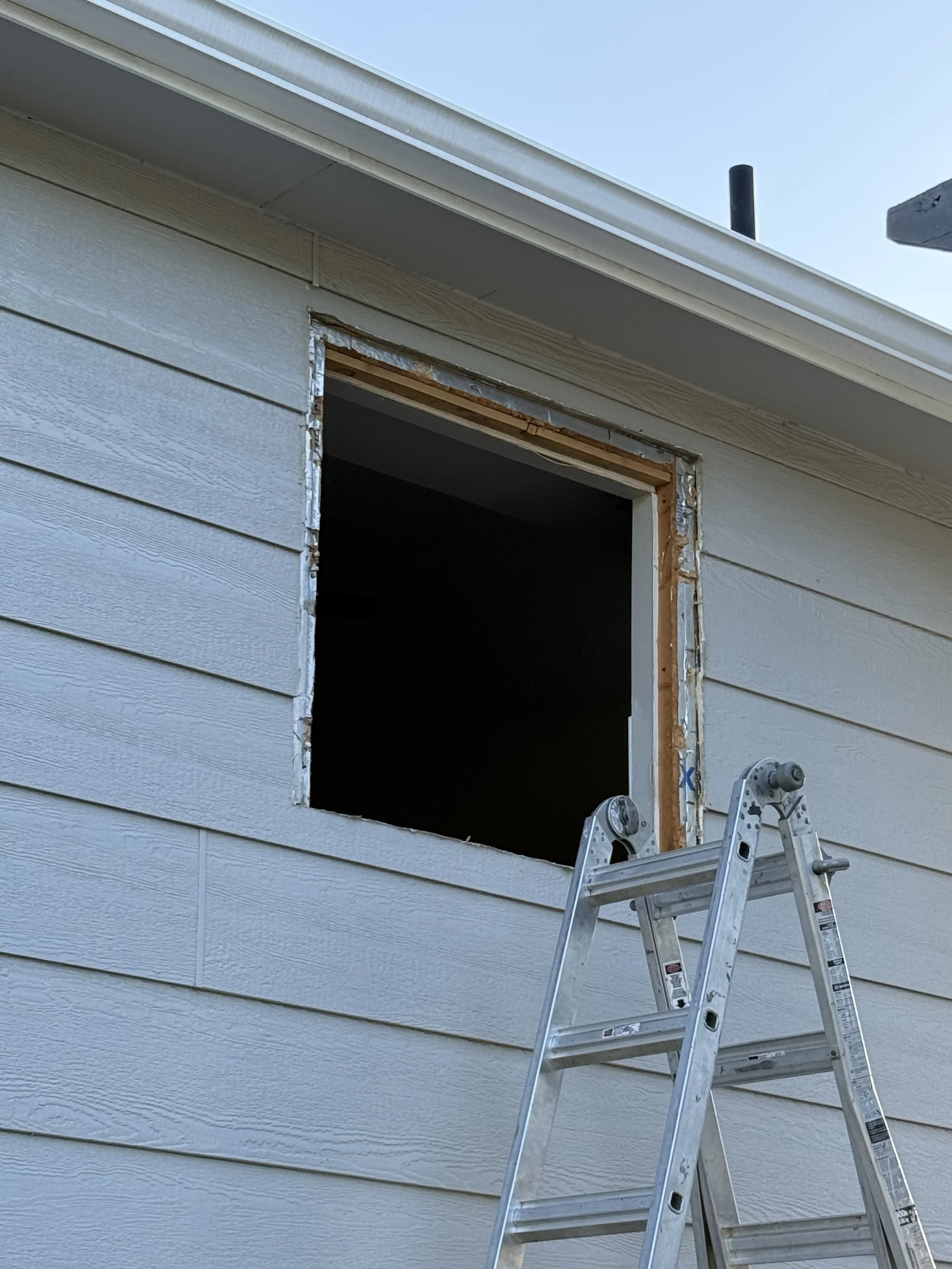 A house exterior wall with beige horizontal siding, an empty window frame, and a ladder positioned in front of the opening.