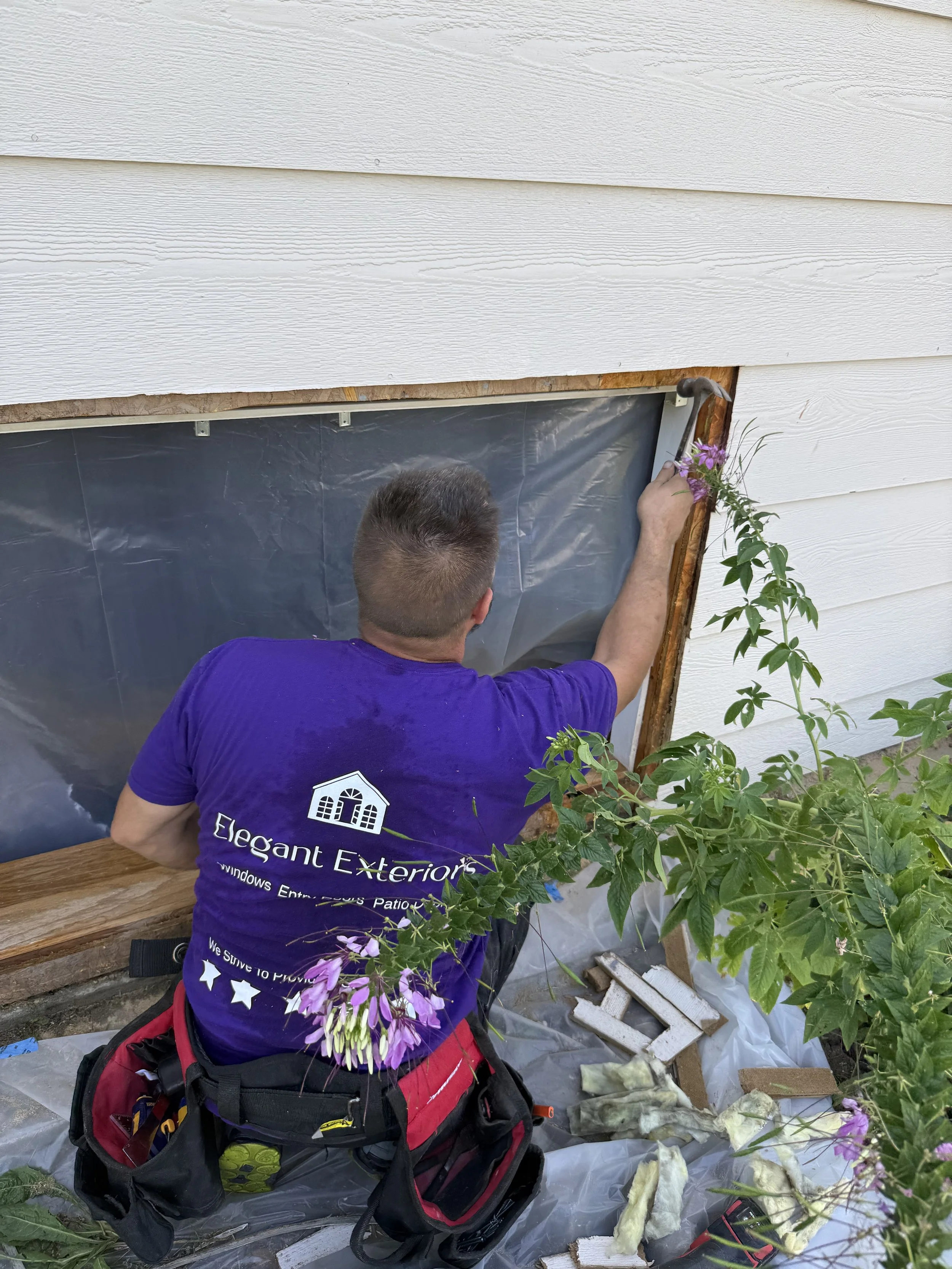 A person installing a window in a house, with tools and debris around, wearing a purple shirt with white text and a backpack.