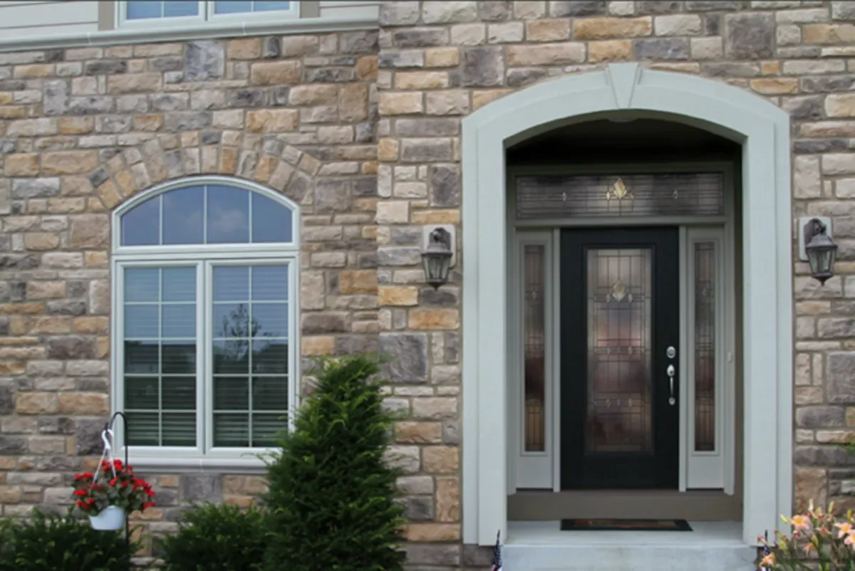 Front exterior of a house with a stone facade, arched window with shutters, black front door with glass panels, and decorative lanterns on either side of the door. There are plants and flowers near the entrance.