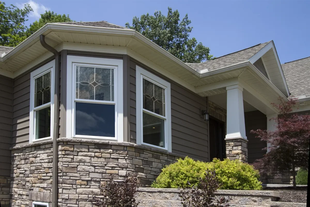 Front view of a house with a stone and siding exterior, bay windows, and a covered porch, surrounded by trees and plants.