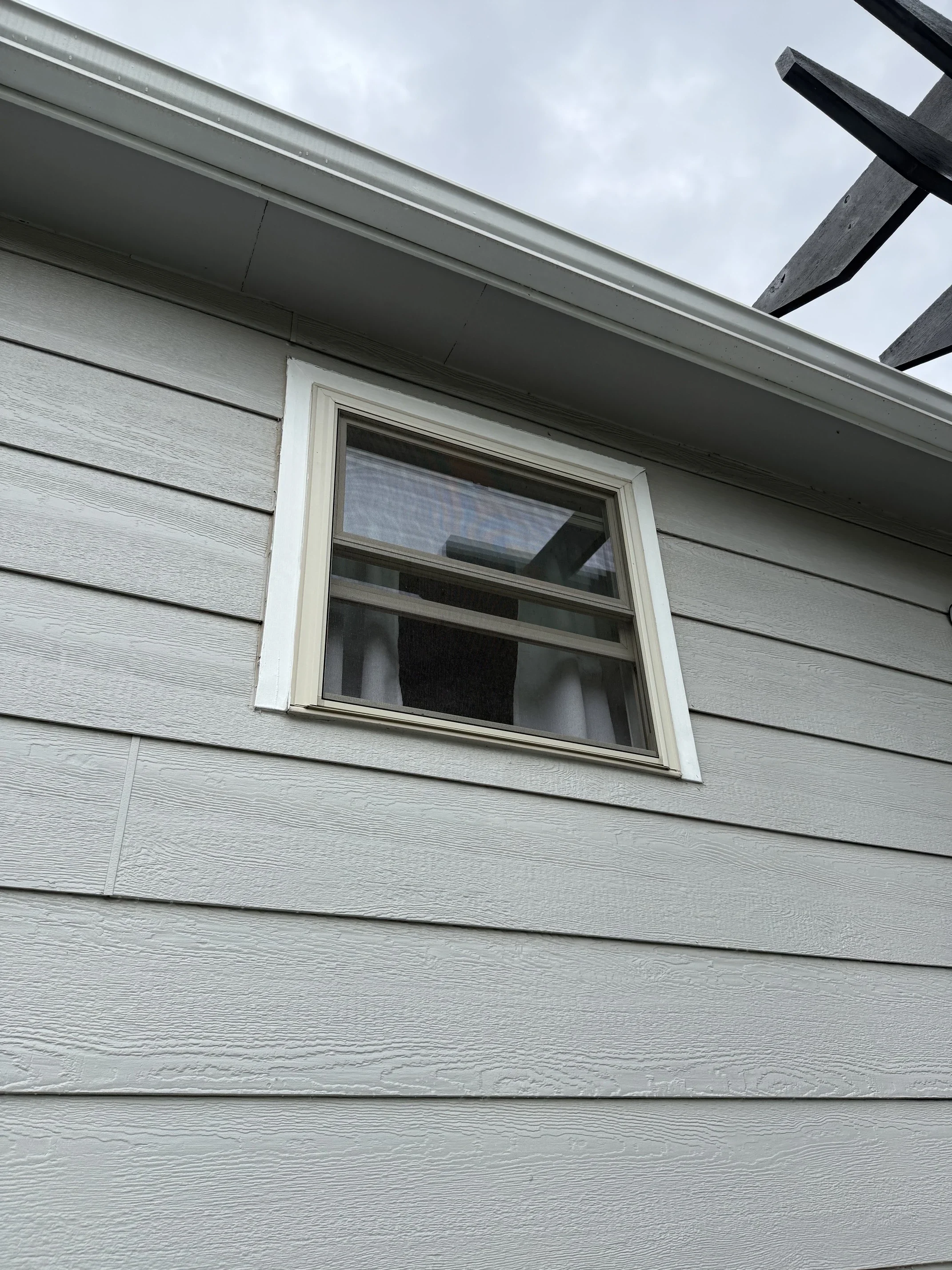 Close-up view of a house's exterior wall with vinyl siding, a window with white trim, an overhanging roof, and parts of a wooden deck or railing, under a cloudy sky.