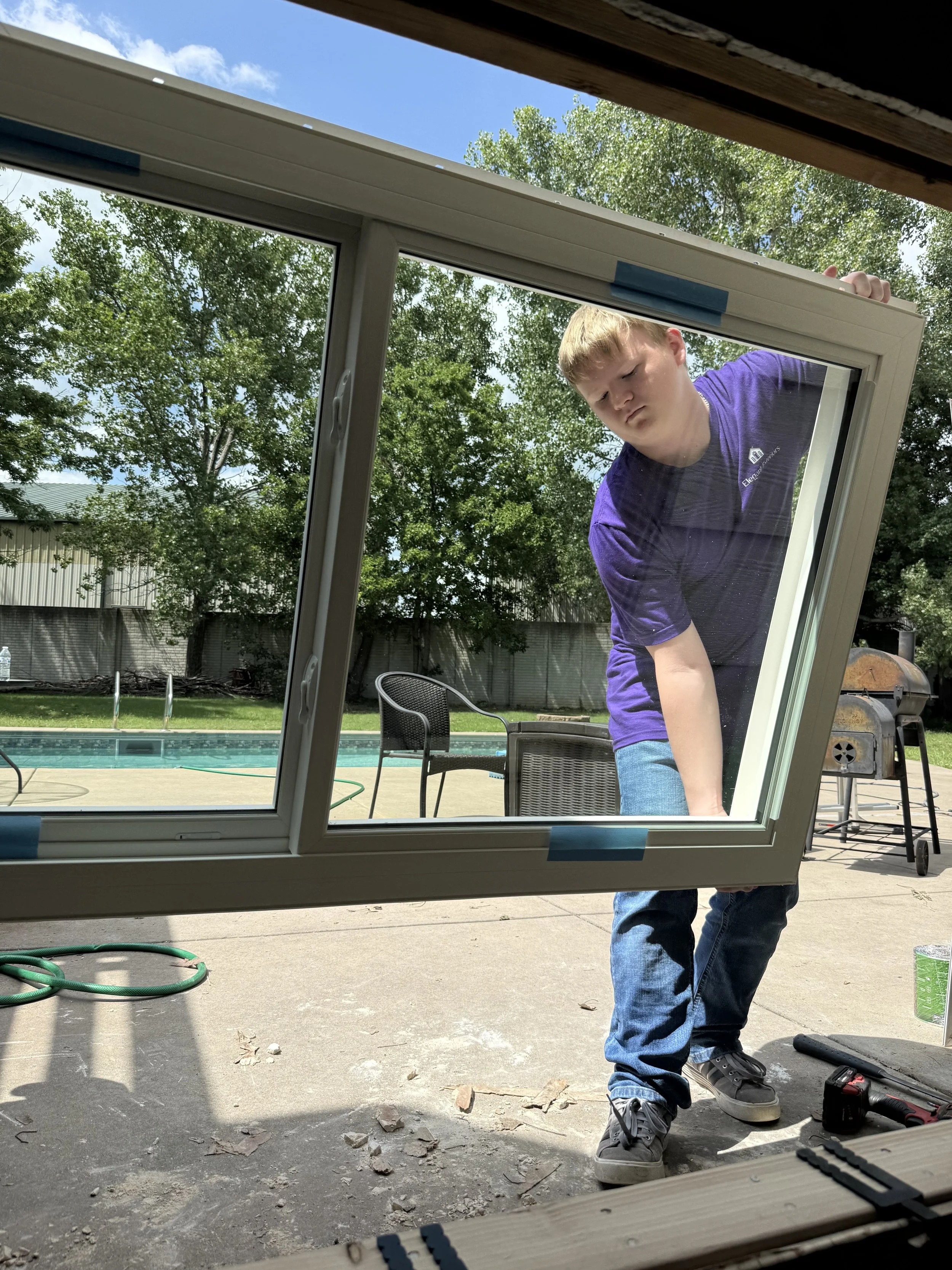 A young man in a purple shirt and jeans is installing a window outdoors. He is leaning over and working on the window frame with tools nearby. The background shows a yard with trees, a swimming pool, and outdoor furniture.