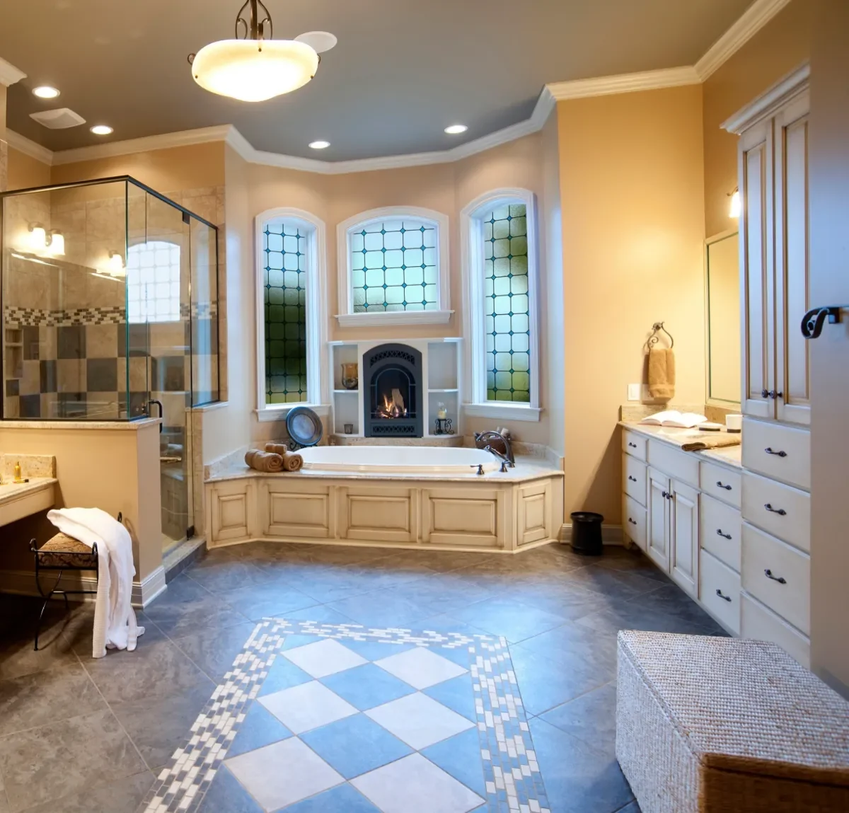 Luxurious bathroom with a bathtub under arched stained glass windows, a fireplace, a separate glass-enclosed shower, and white cabinetry.