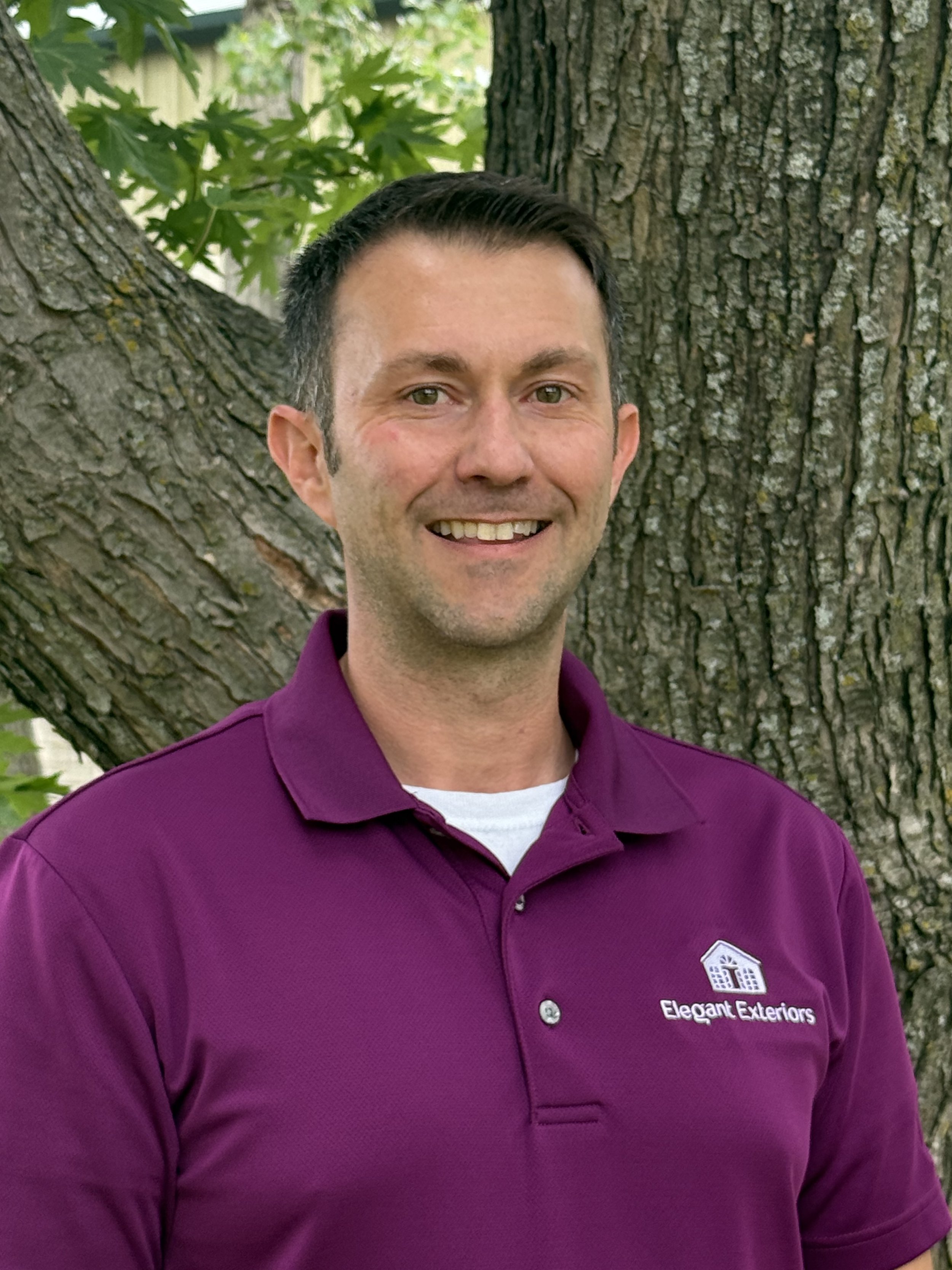 Chris Schmitz the Owner smiling outdoors in front of a tree, wearing a purple polo shirt with the logo and text 'Elegant Exteriors'.