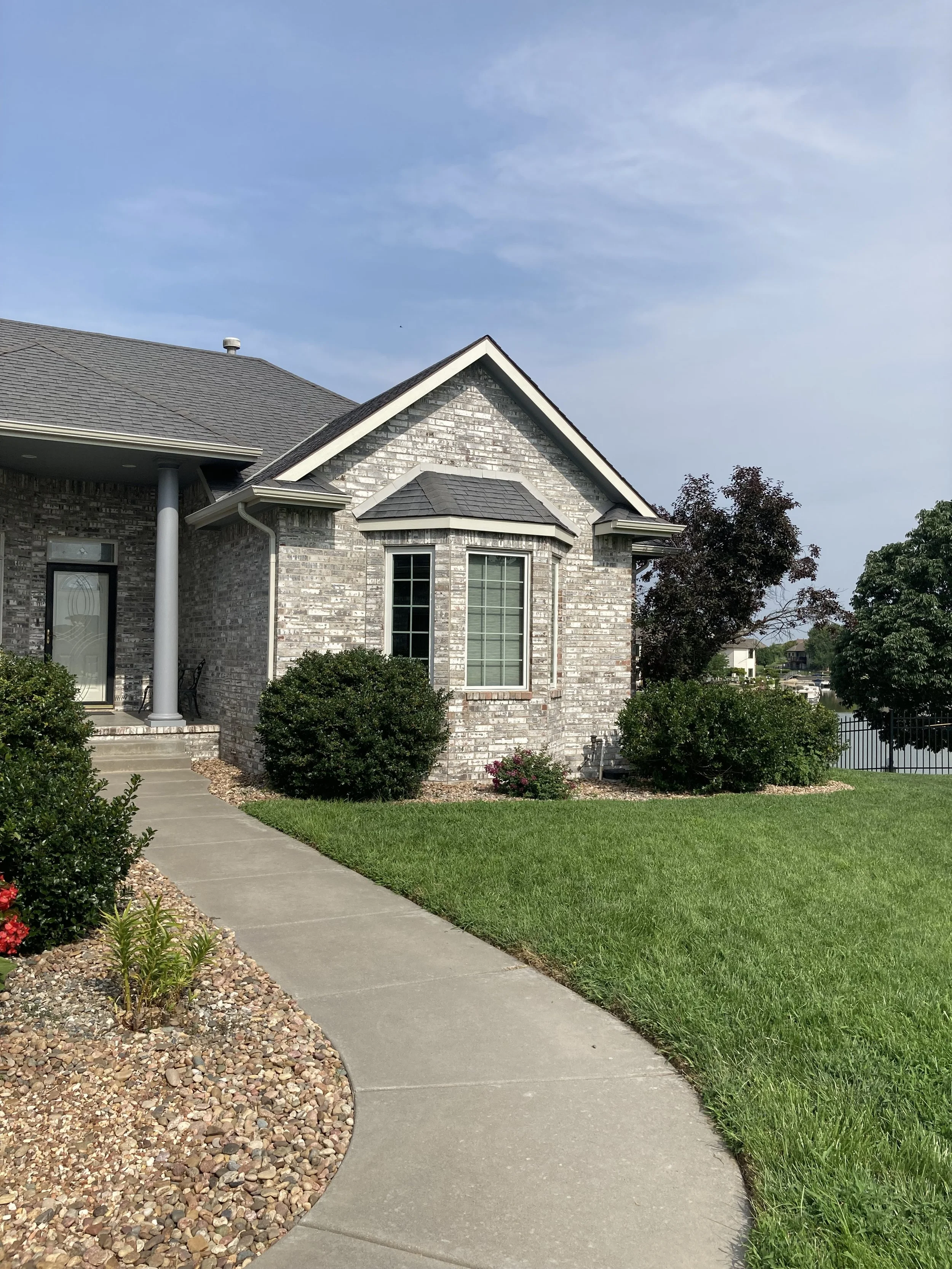 Front yard and house with brick exterior, gray roof, large window, walkway, bushes, and trees under a blue sky.