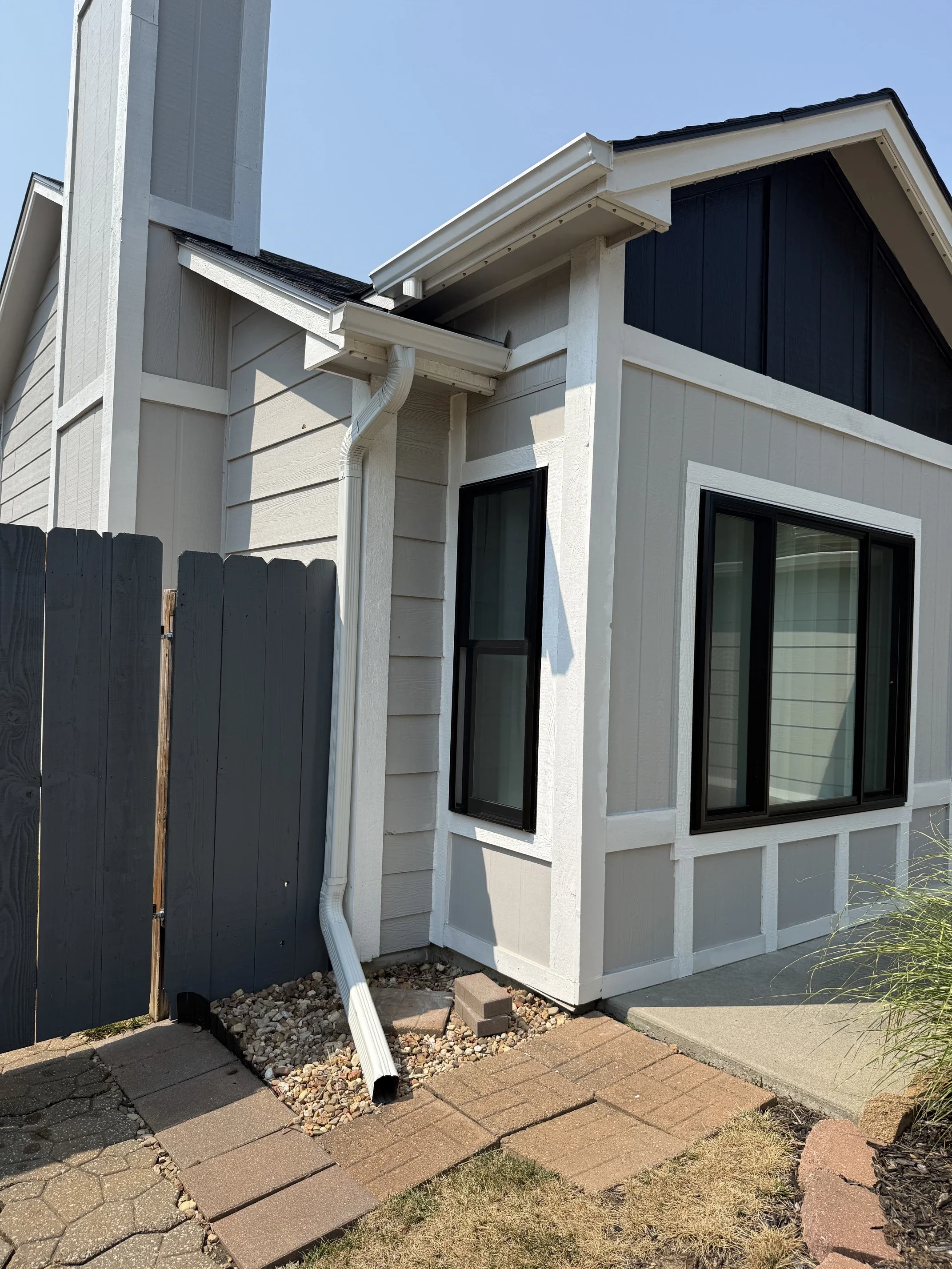 A house exterior showing a corner with siding, window, and roof with a gutter. There is a wooden fence on the left and a small garden area with stones and paving stones in front of the house.