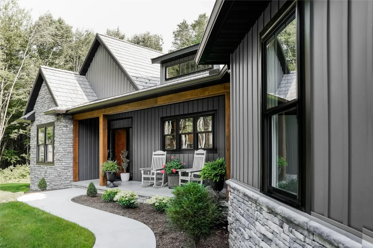 Modern house with black metal siding, stone accents, and a small front porch with chairs and potted plants, surrounded by green grass and trees.