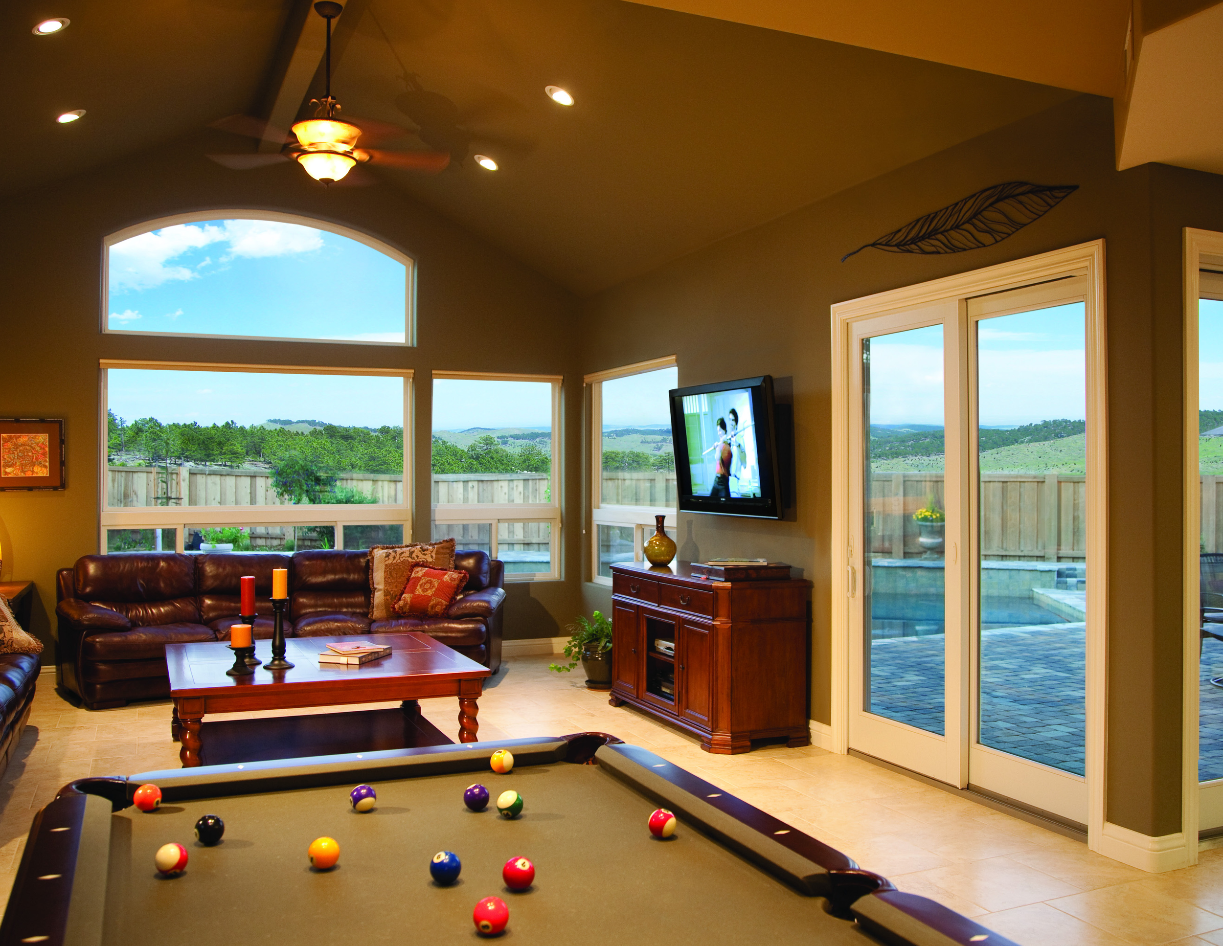 A living room with a pool table in the foreground, a leather sofa, a wooden sideboard, and a flat-screen TV on the wall. Large windows and sliding glass doors reveal a backyard with a fence and green hills. There is a ceiling fan and recessed lighting, and a decorative metal leaf on the wall.