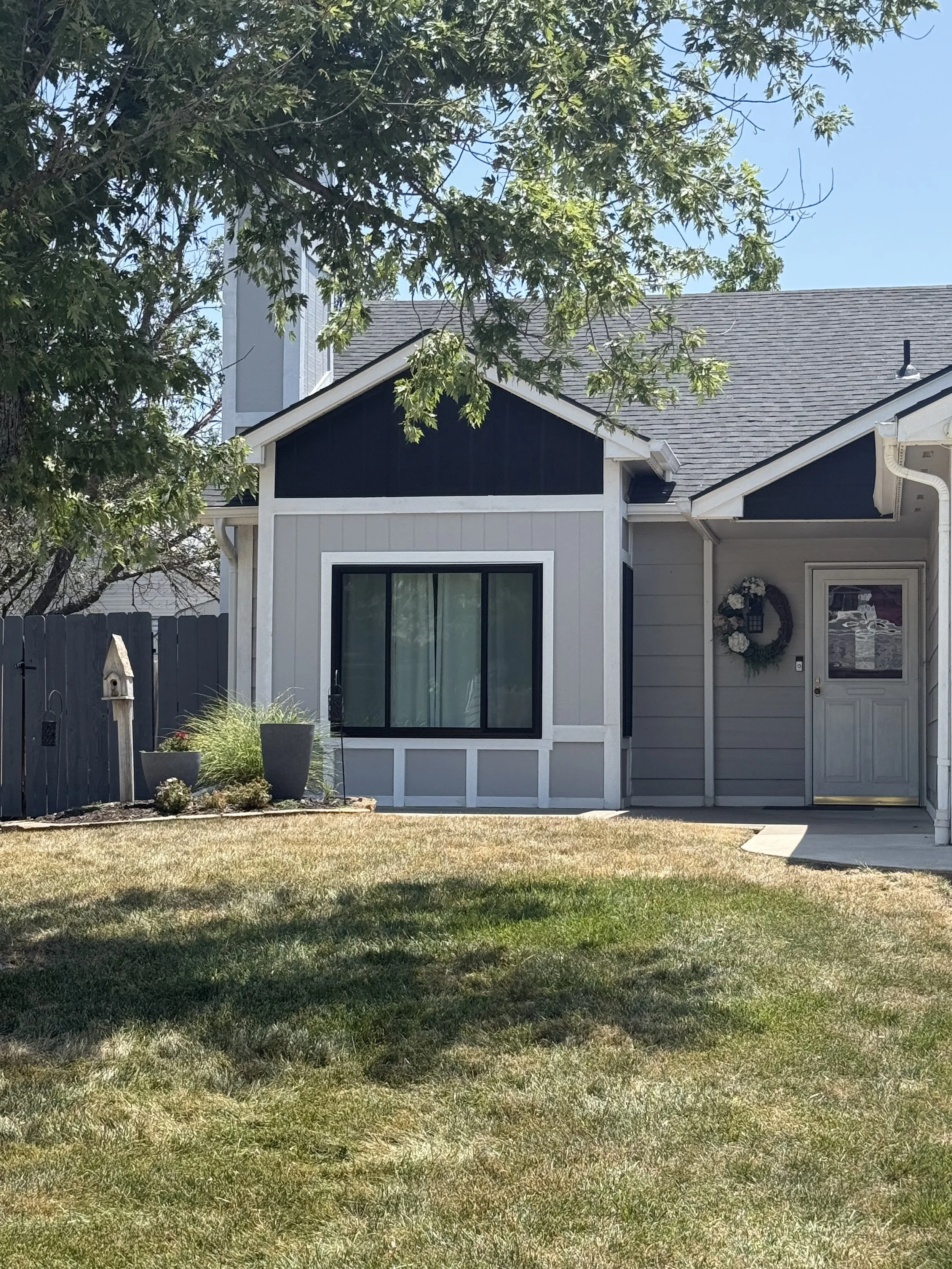 The photo shows the front of a house with beige and navy blue exterior, a large window, a front door with a wreath, and a small lawn with a patch of grass in the foreground. There is a tree with green leaves partially blocking the view at the top.