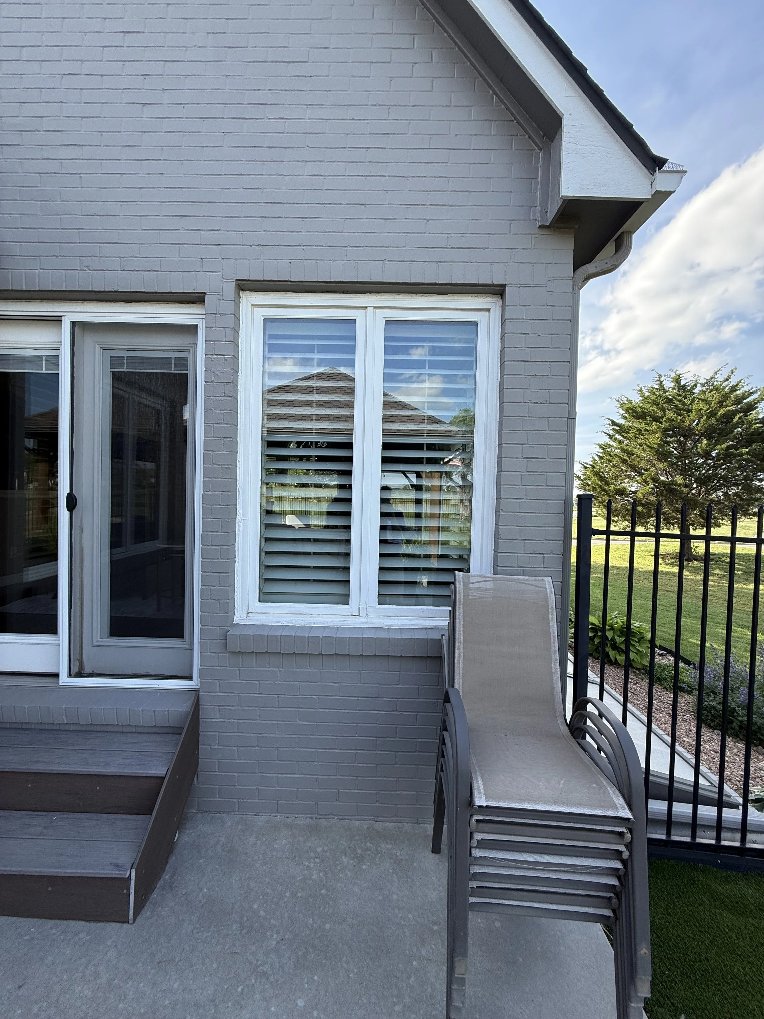 Backyard patio area with stacked chairs, a sliding glass door, windows with shutters, a white brick house exterior, and a black metal fence with some greenery and a tree outside.