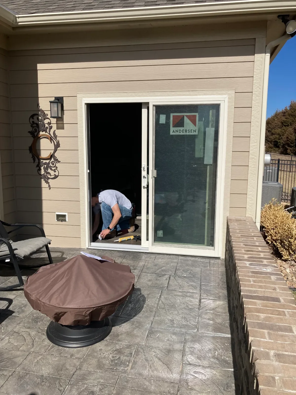A person installing or repairing a sliding glass door on a house exterior, with outdoor furniture on the patio and a brick border.
