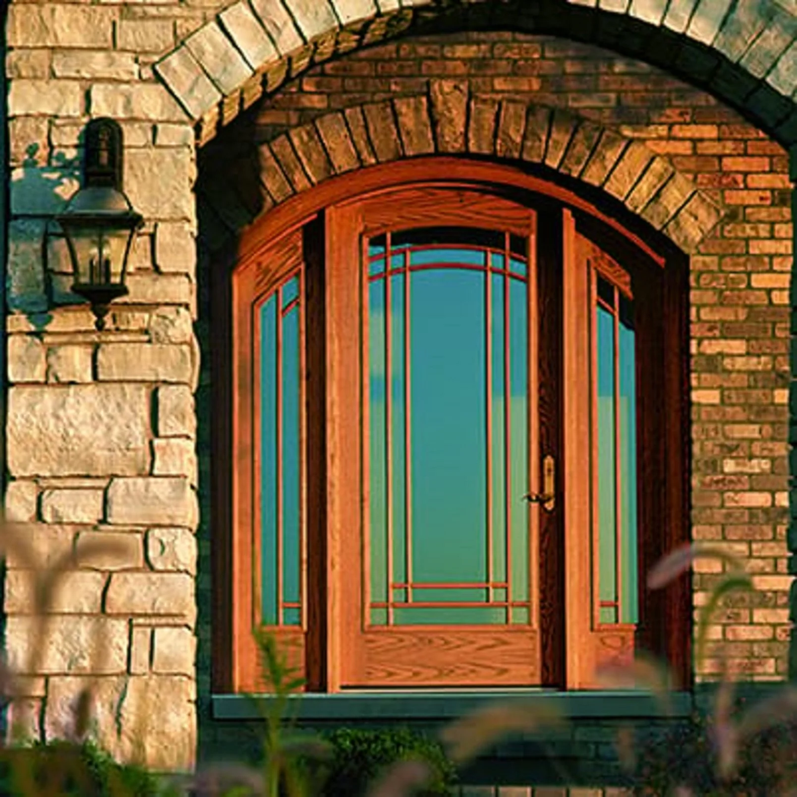 A large wooden door with glass panels in a brick wall archway, flanked by an exterior stone wall light fixture.
