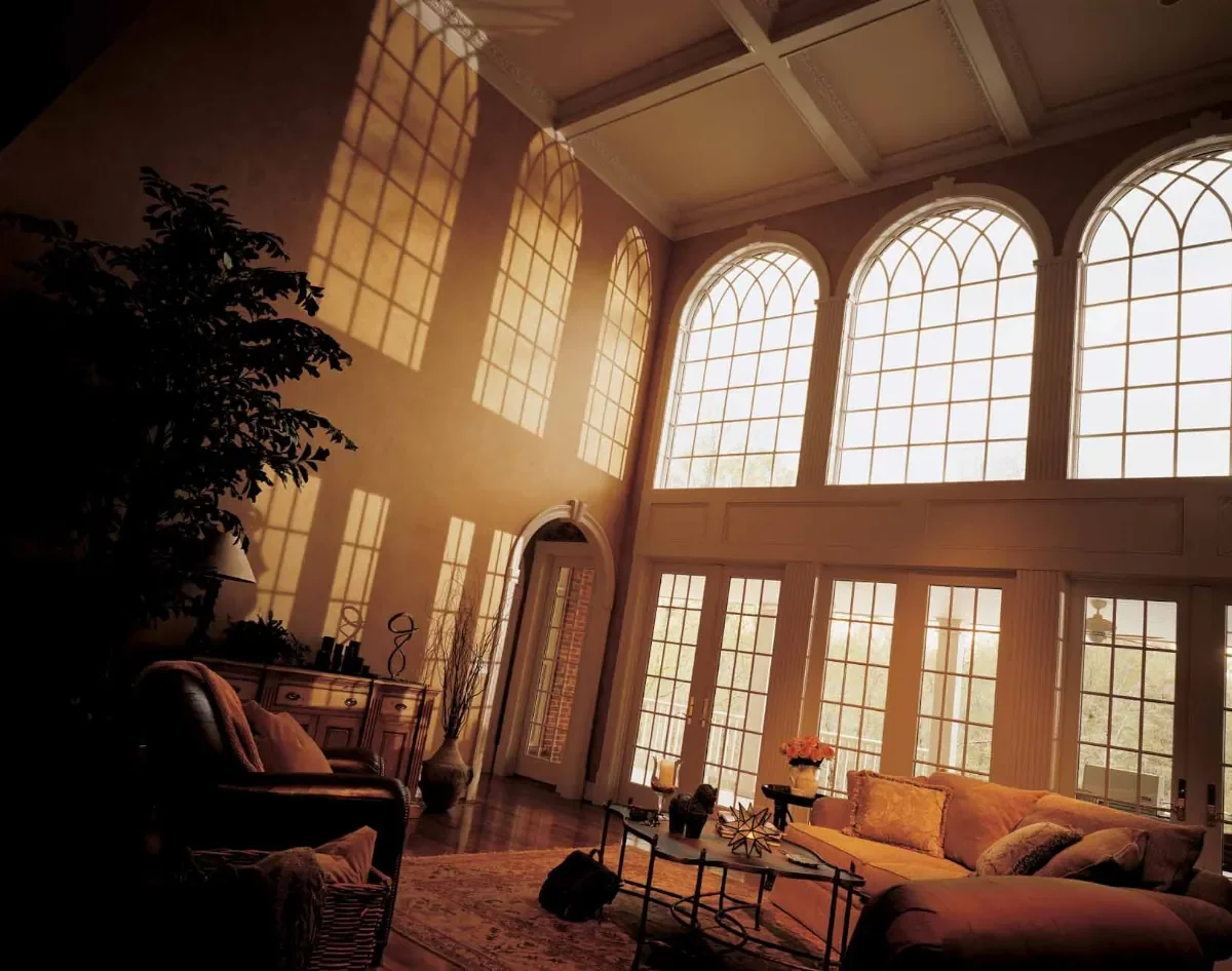 Sunlight streaming through large arched and rectangular windows into a spacious, warmly lit living room with a yellow sofa, a wooden coffee table, and decorative items.