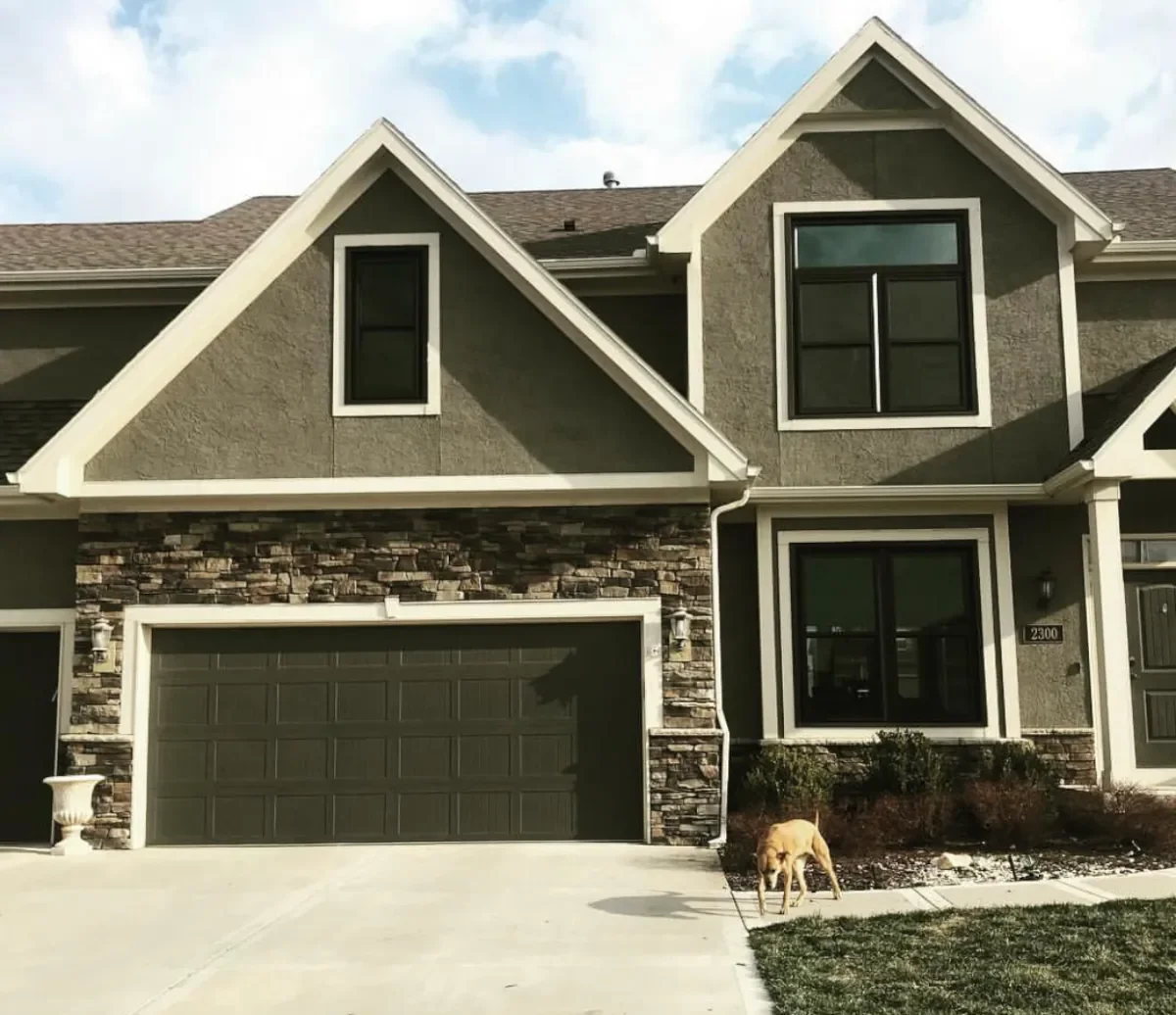 Two-story house with gray walls, stone accents, and dark windows. A dog is sniffing near the shrubs in the front yard.