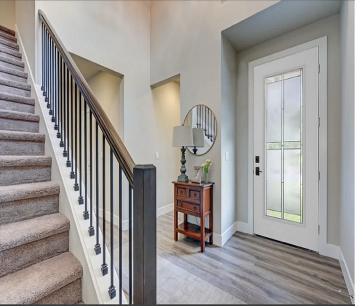 Entryway with front door, small wooden side table, lamp, round mirror, and staircase with beige carpet and black metal railing.