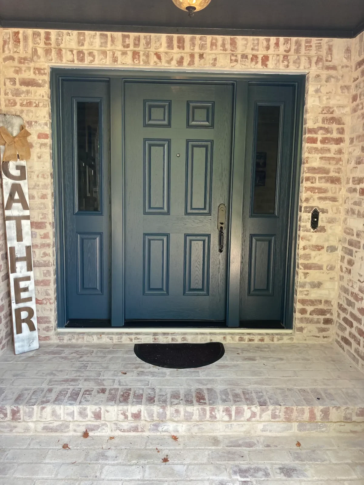A front porch entrance with a dark green front door flanked by two narrow windows. The porch has a brick wall exterior and a small black mat in front of the door.