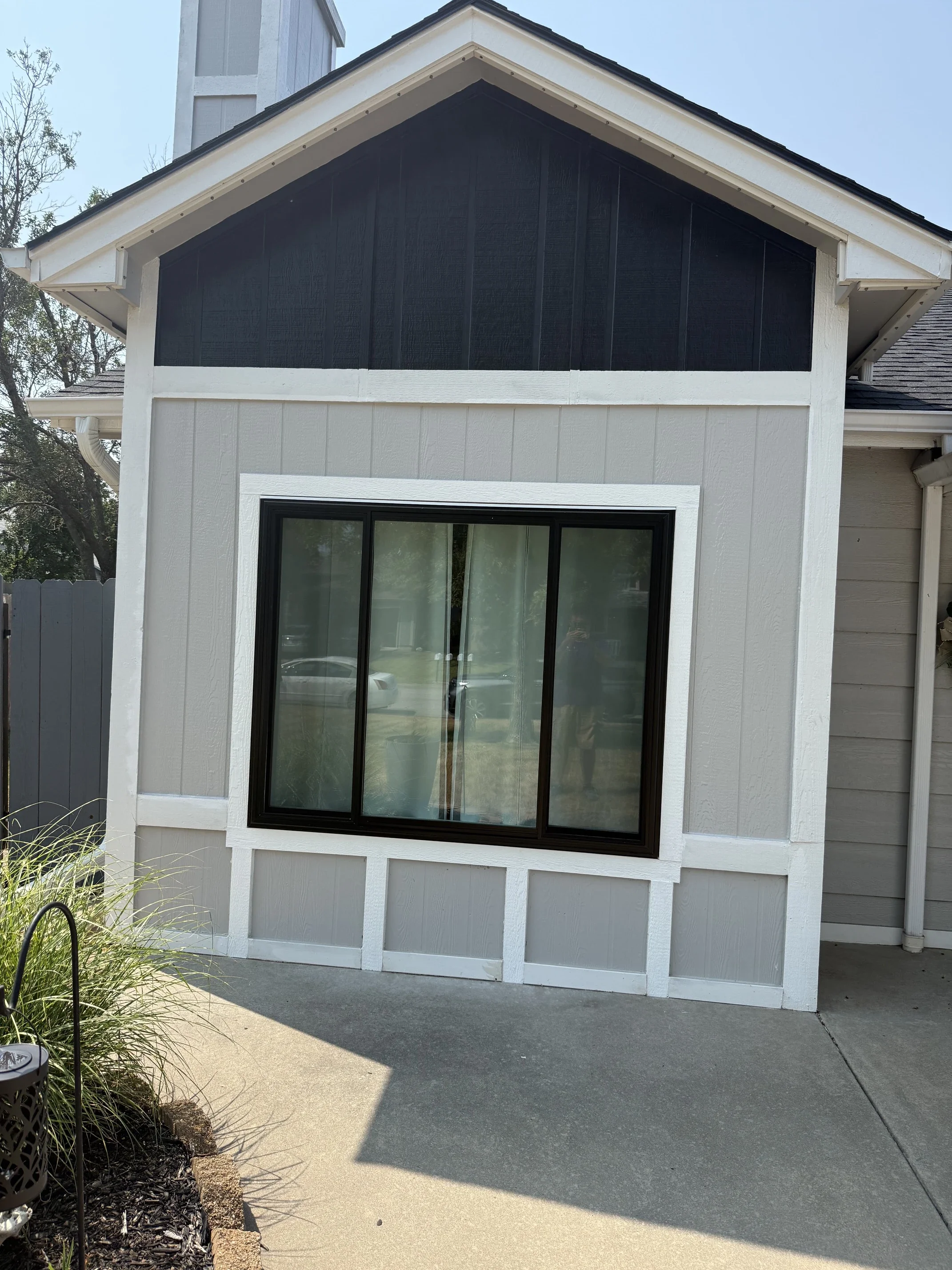 The back of a house with large black framed sliding glass doors and a window, beige siding, and an upper section with black wood paneling. There is a concrete patio and some landscaping with a garden bed and a metal plant holder.
