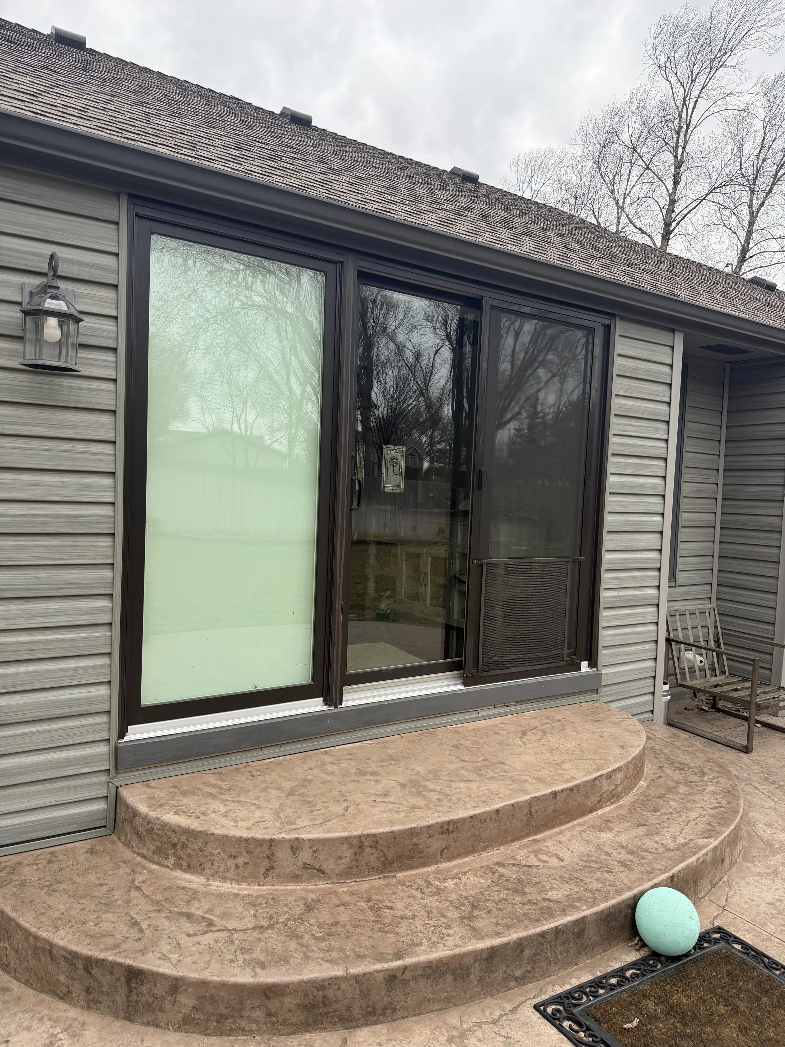 Backyard scene featuring a set of three curved wooden steps leading up to a sliding glass door with brick-colored flooring. There is a small outdoor mat, a decorative ball, and a bench on the right. The house has beige vinyl siding, a black-framed sliding door, and a wall-mounted lantern light. Bare trees and an overcast sky are visible in the background.