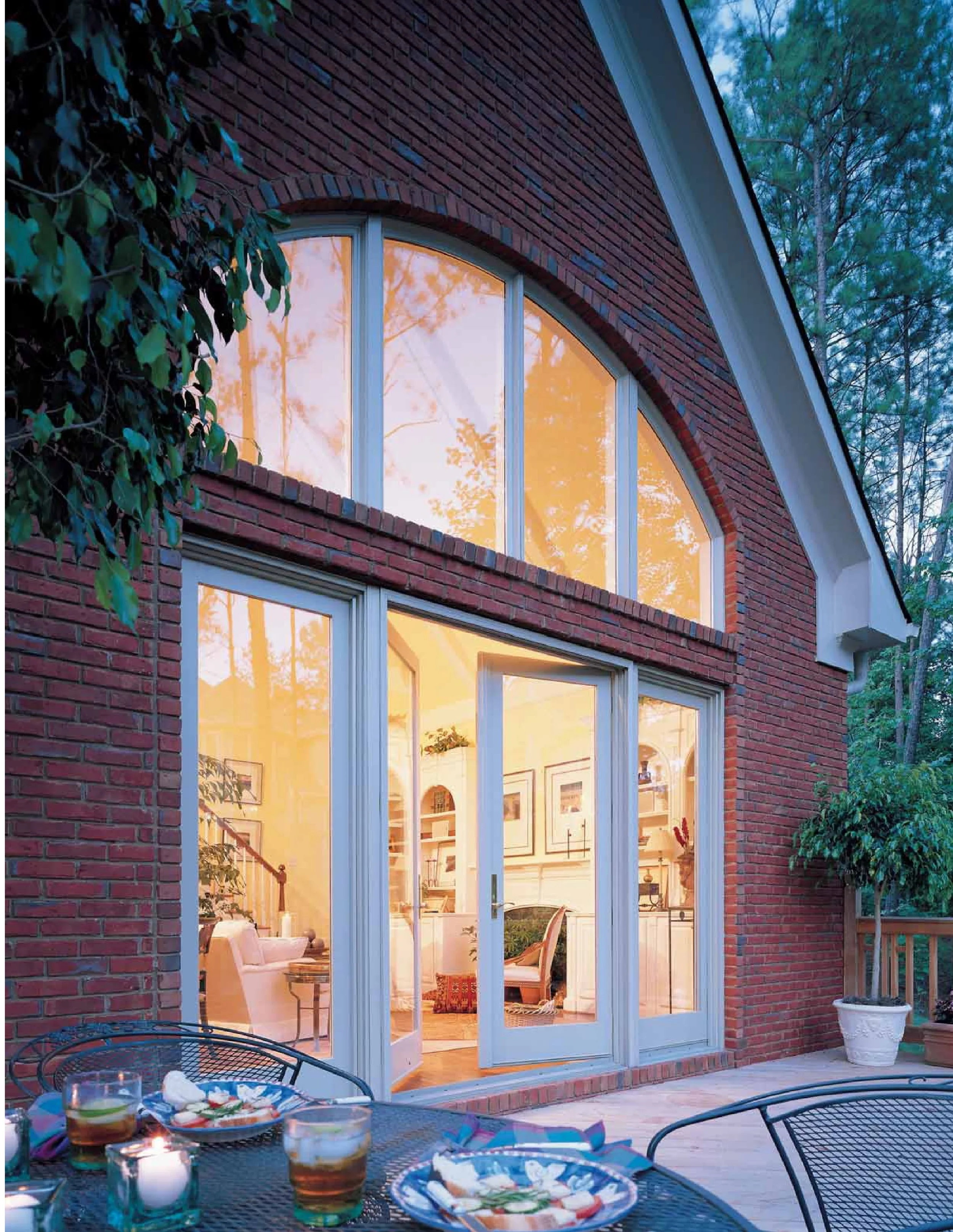 View of a house with a brick exterior and large arched window, illuminated from inside, evening setting, outdoor patio with table and drinks.