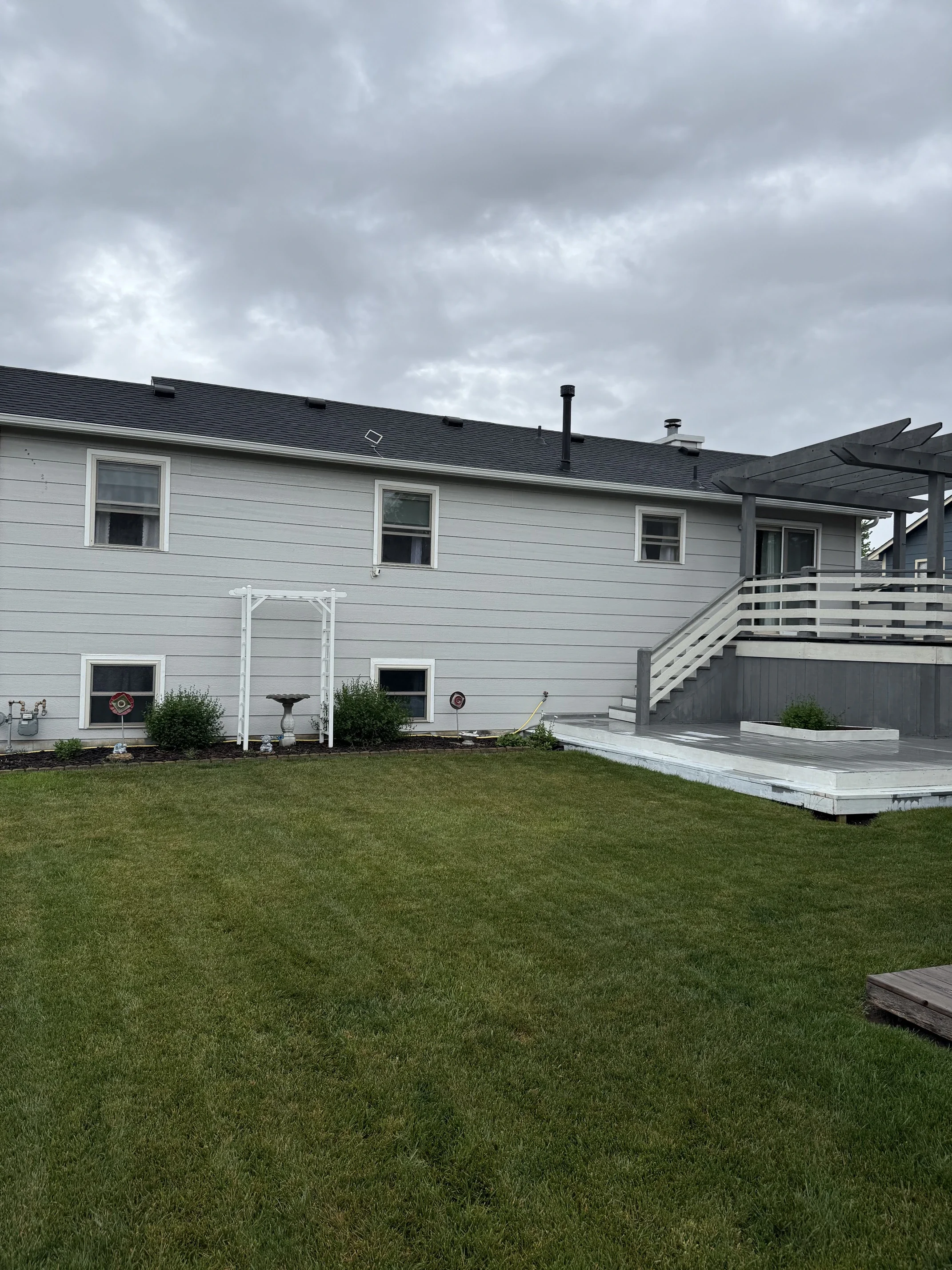 Backyard with a white house, green lawn, potted plants, bird bath, and cloudy sky.