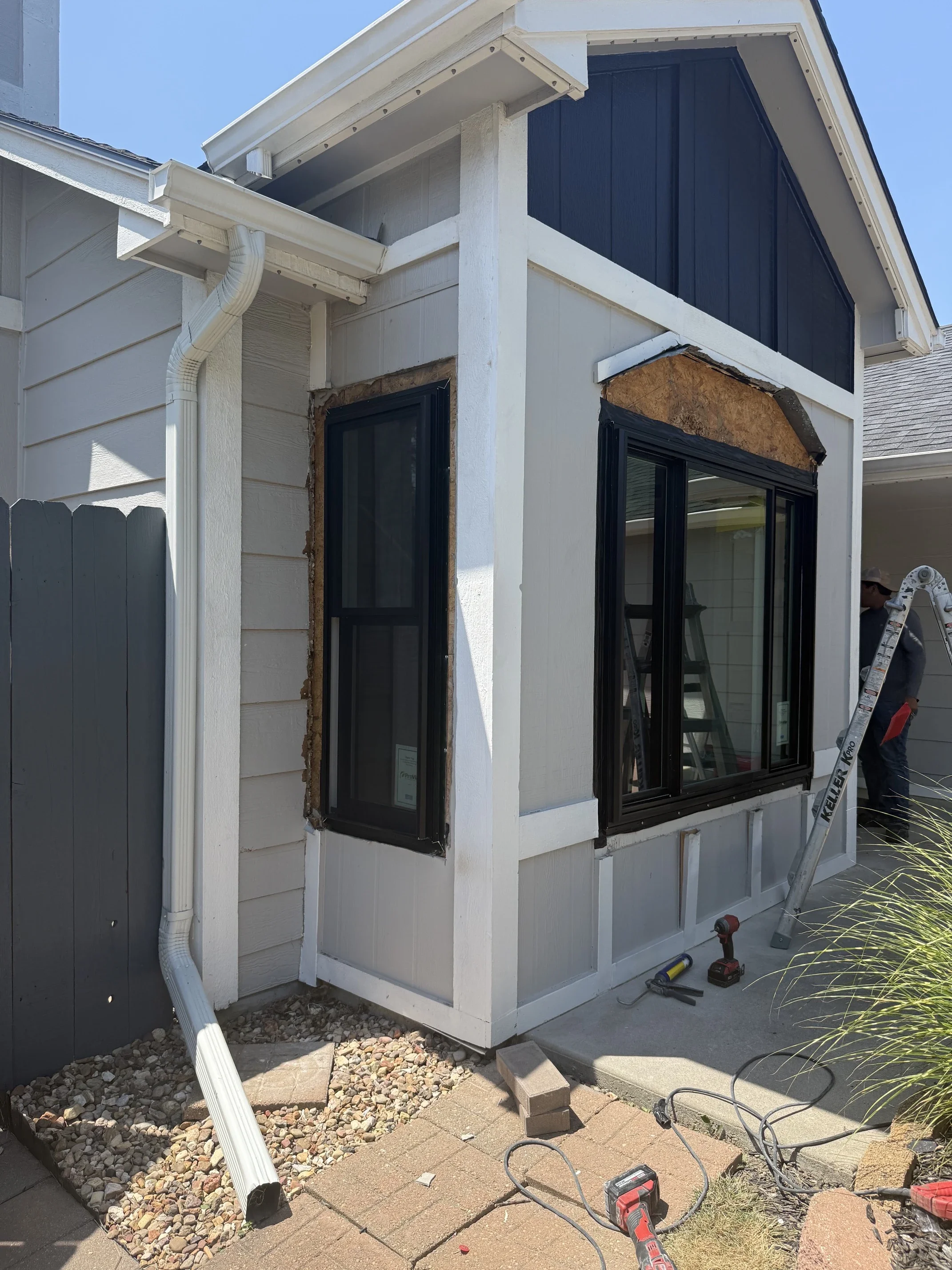 Construction work on a house exterior, with tools and materials on the ground, including a drill, ladder, and bricks. A person is visible in the background working near the house with a ladder.