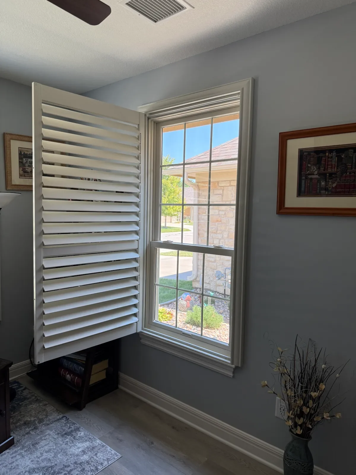 A window with white plantation shutters partially open, showing a view of a stone house and a garden outside, with framed pictures on the wall in a light blue room.