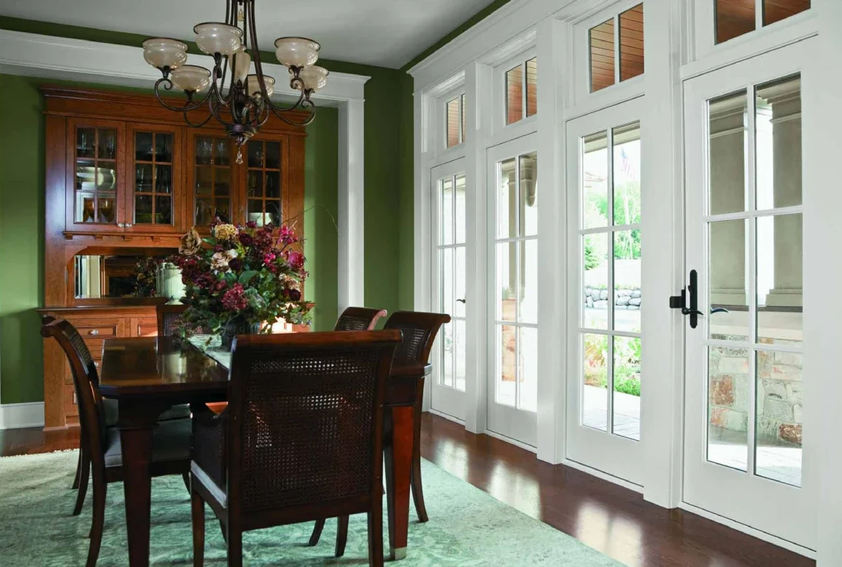 Dining room with green walls, dark wood dining table and chairs, a large floral centerpiece, a chandelier, and multiple white-paneled glass doors leading outside.